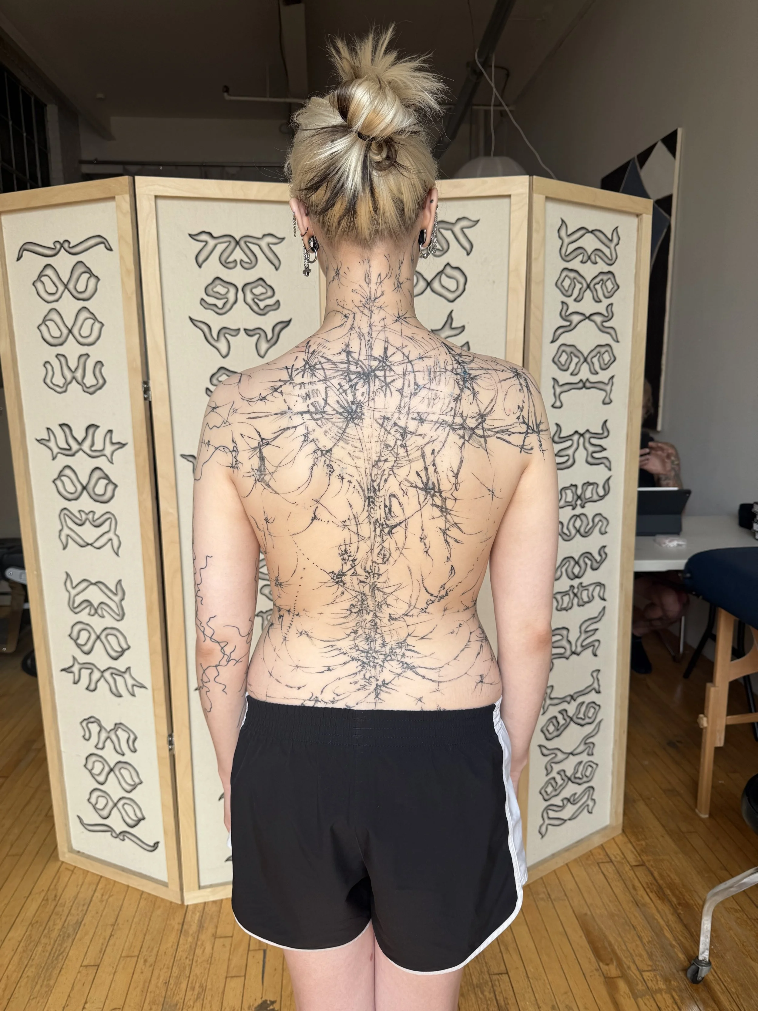 Back of a woman with blonde hair in a messy bun, featuring extensive black body tattoos that resemble interconnected thorny vines covering her back, shoulders, and arms. She is standing in front of a decorative room divider with abstract black and gr