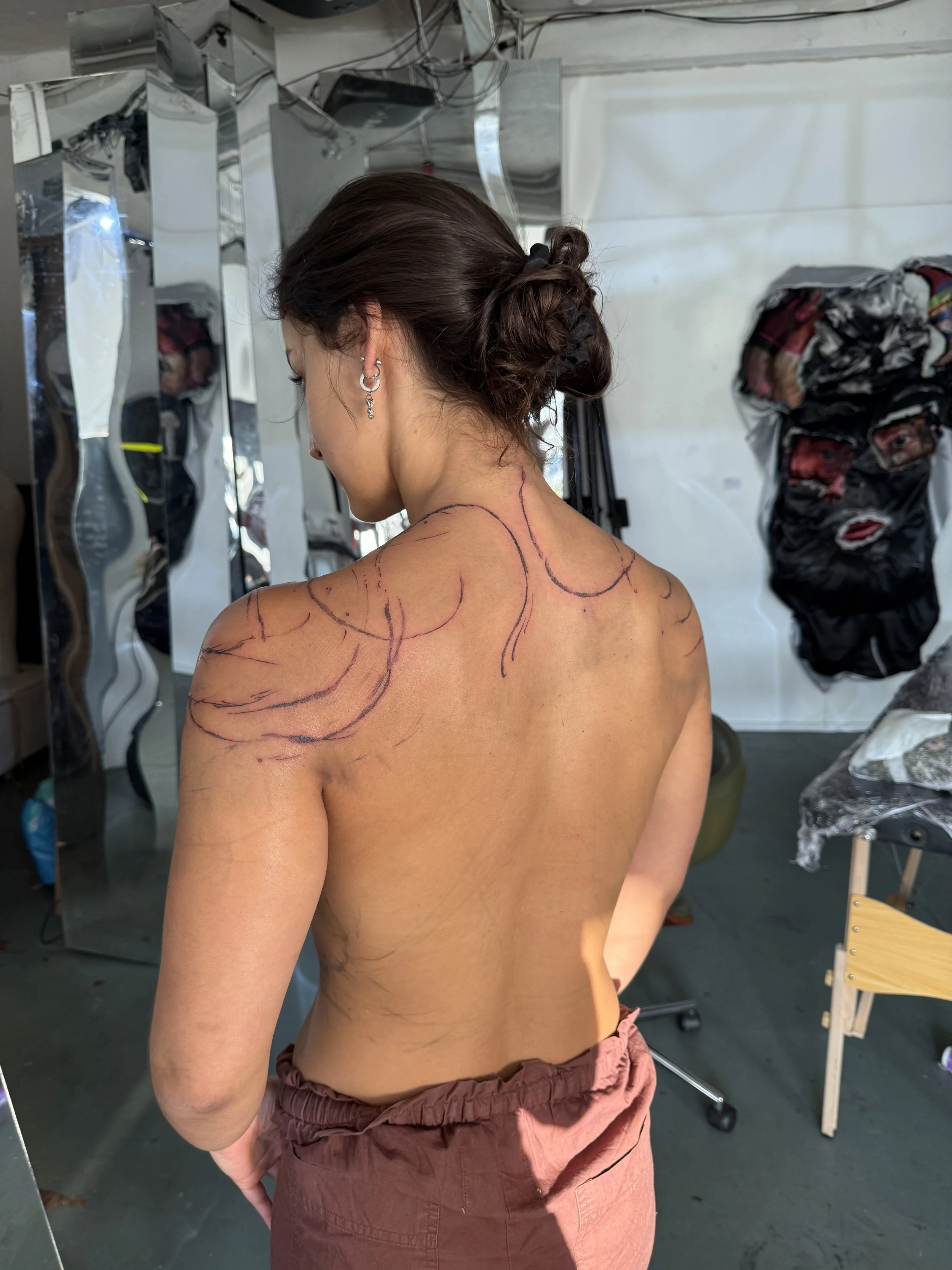 Woman with dark brown hair in a bun, wearing earrings, seen from behind, with markings on her back and shoulder in a studio or makeup room.