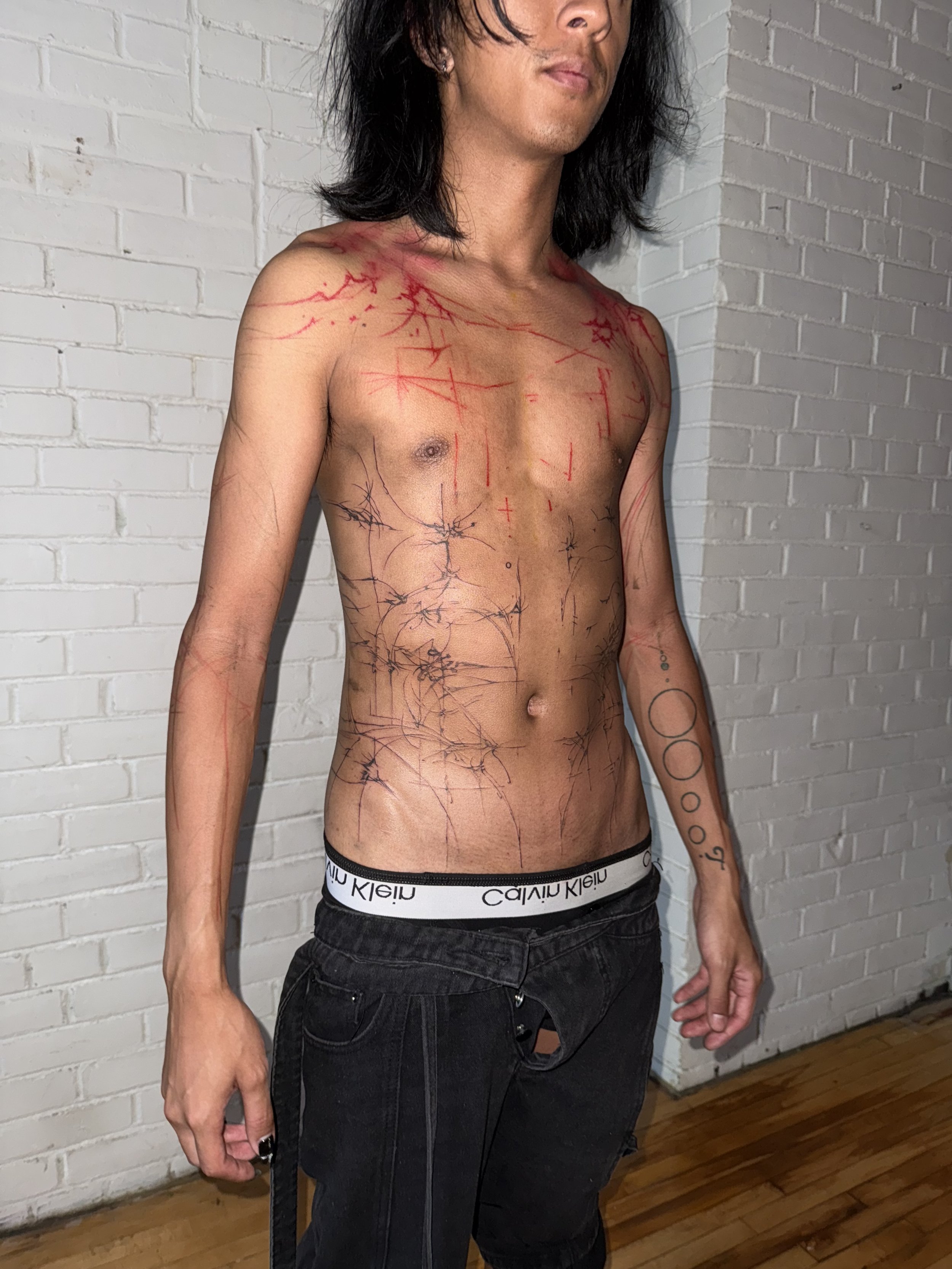 A shirtless man with long dark hair and tattoos on his chest, arms, and torso standing against a white brick wall.