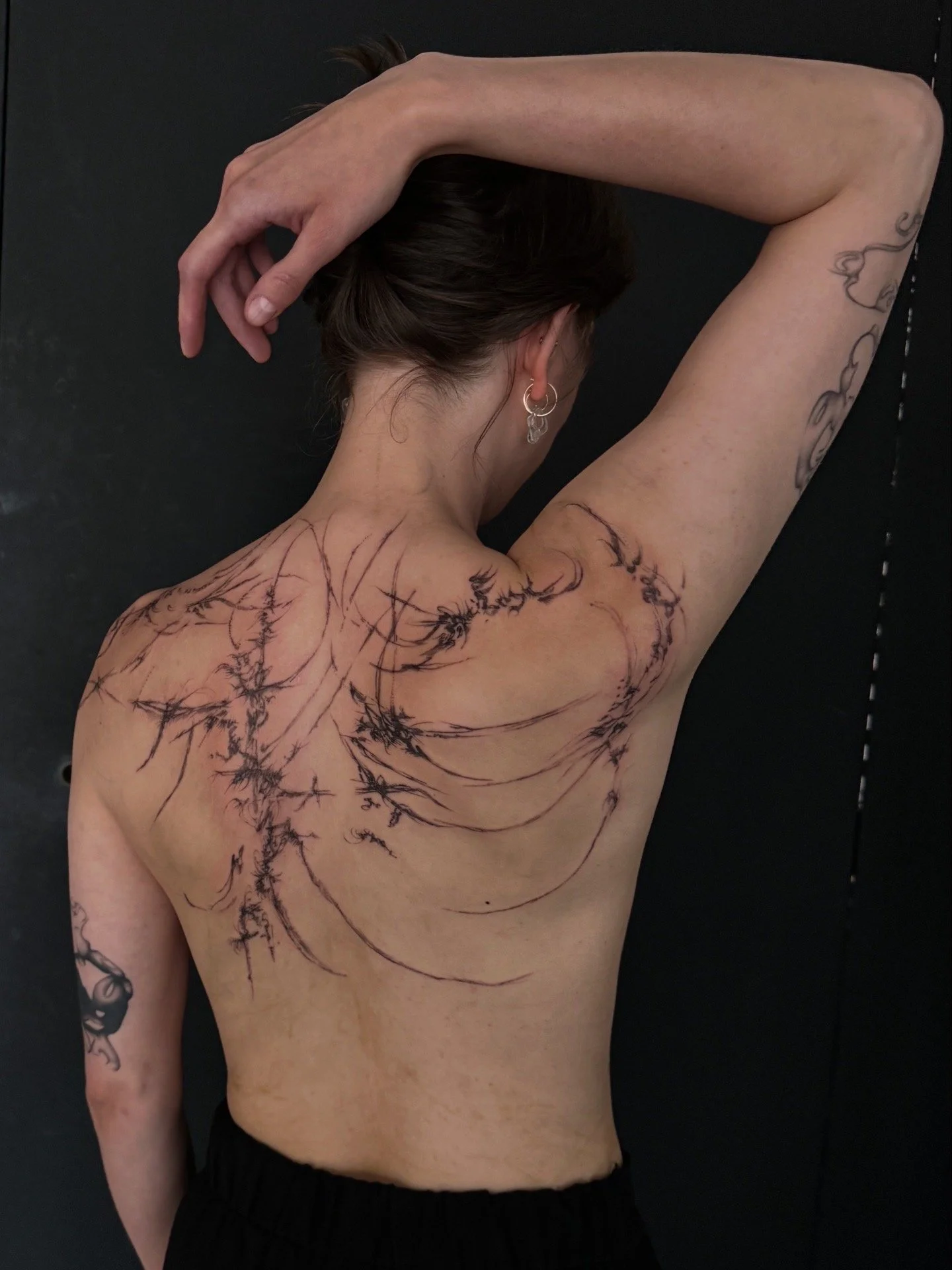A woman with dark hair in a bun, earrings, and tattoos on her back, shoulder, and arm, poses with her back to the camera against a black background, her right arm raised and hand resting on her head, exposing her tattooed back and armpit.