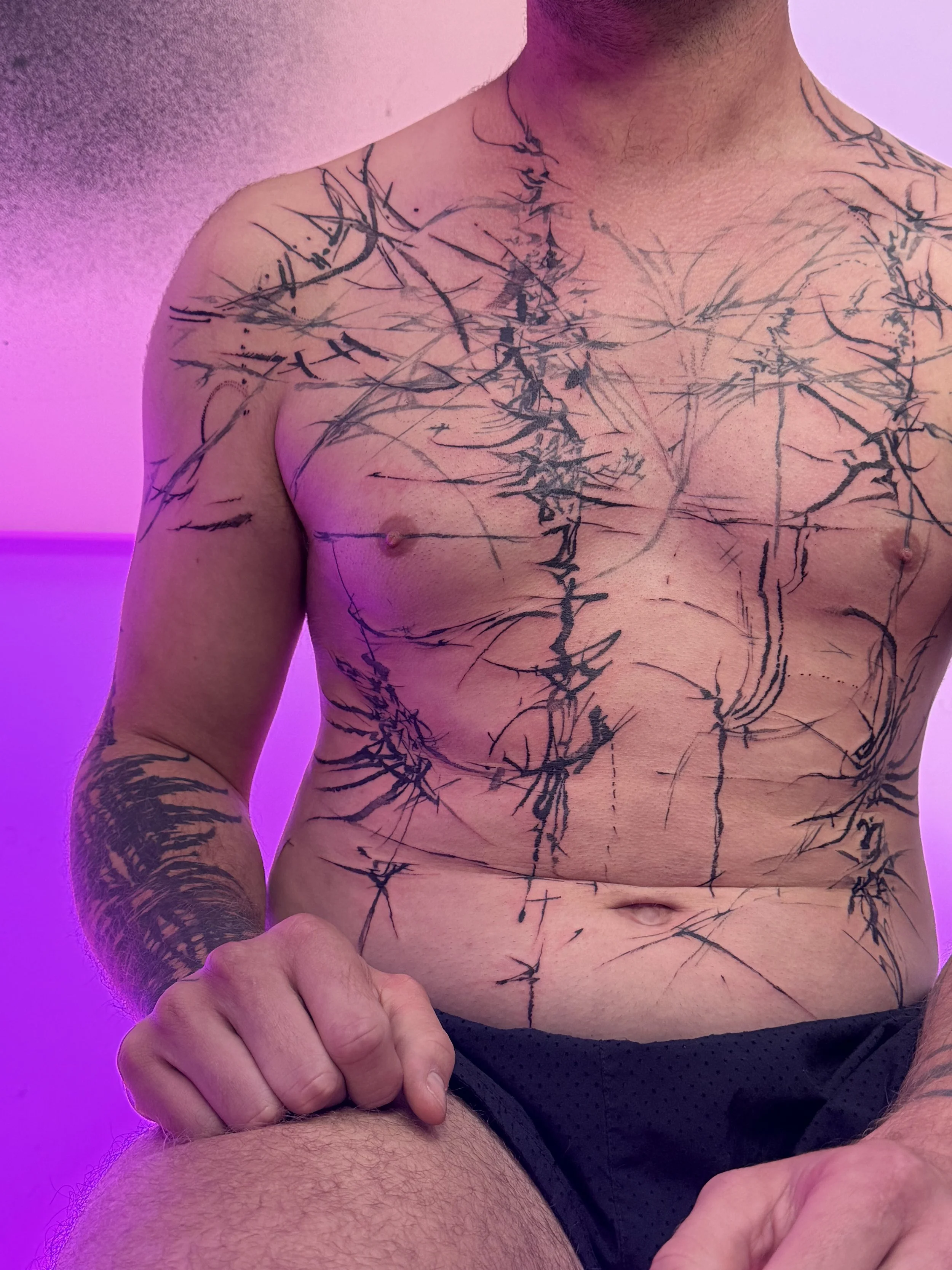 A shirtless man with black tattoos on his chest, arms, and shoulders, sitting against a purple-lit background.
