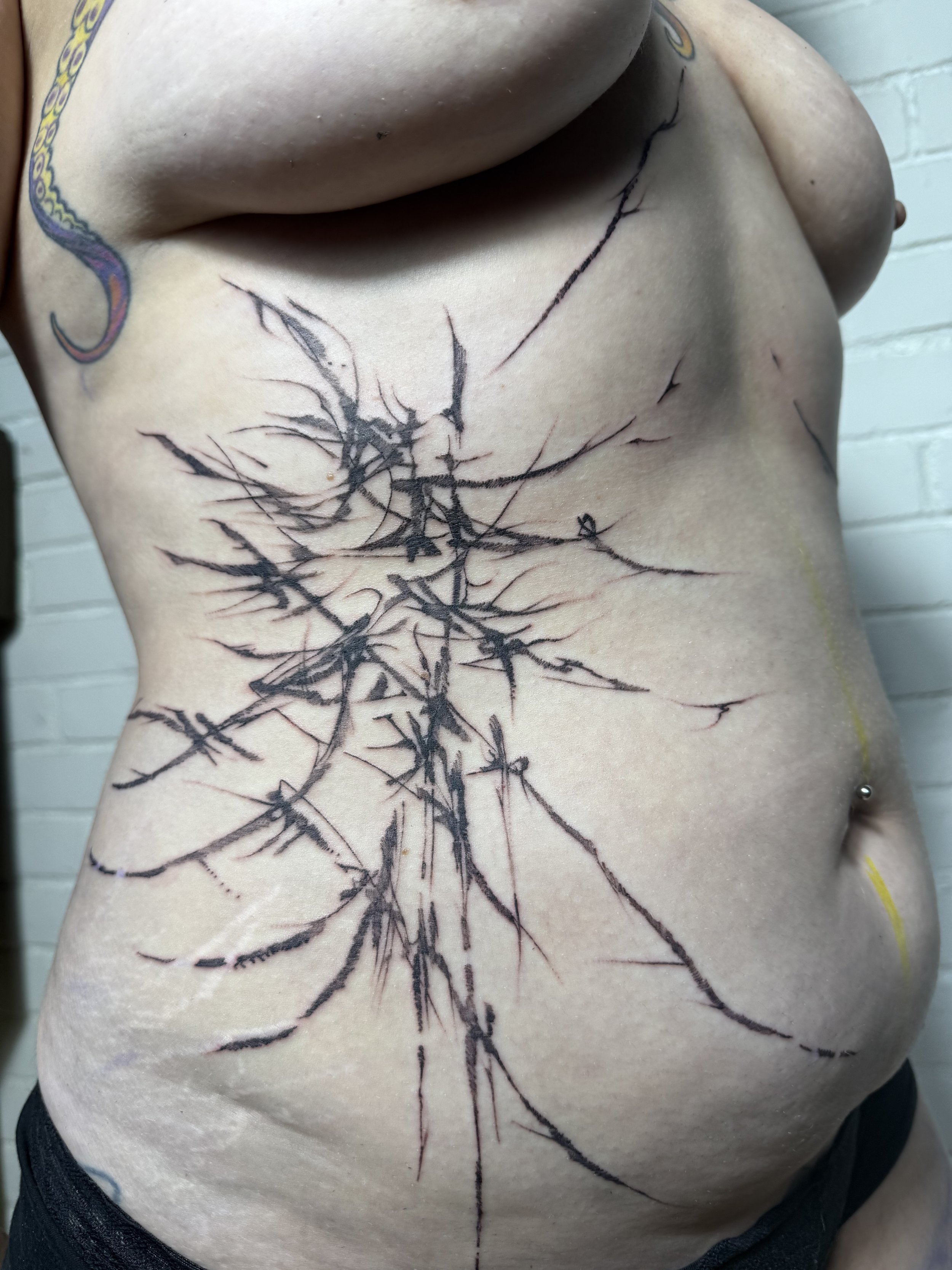 A person's torso with a large, abstract black tattoo resembling a tree or branches on the right side of the chest and abdomen.