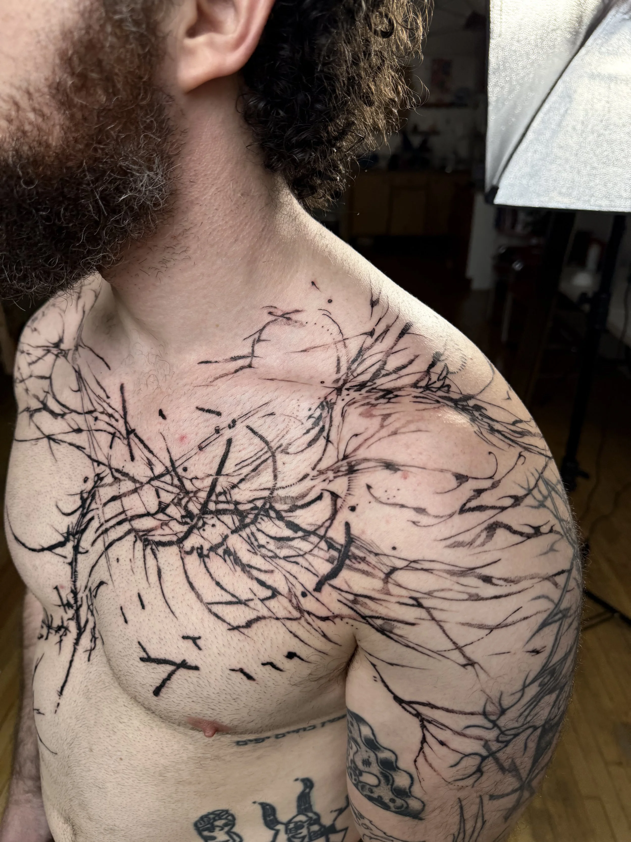 A man with a beard and curly hair showing a large black ink tattoo on his upper chest and shoulder, with abstract branch-like lines.
