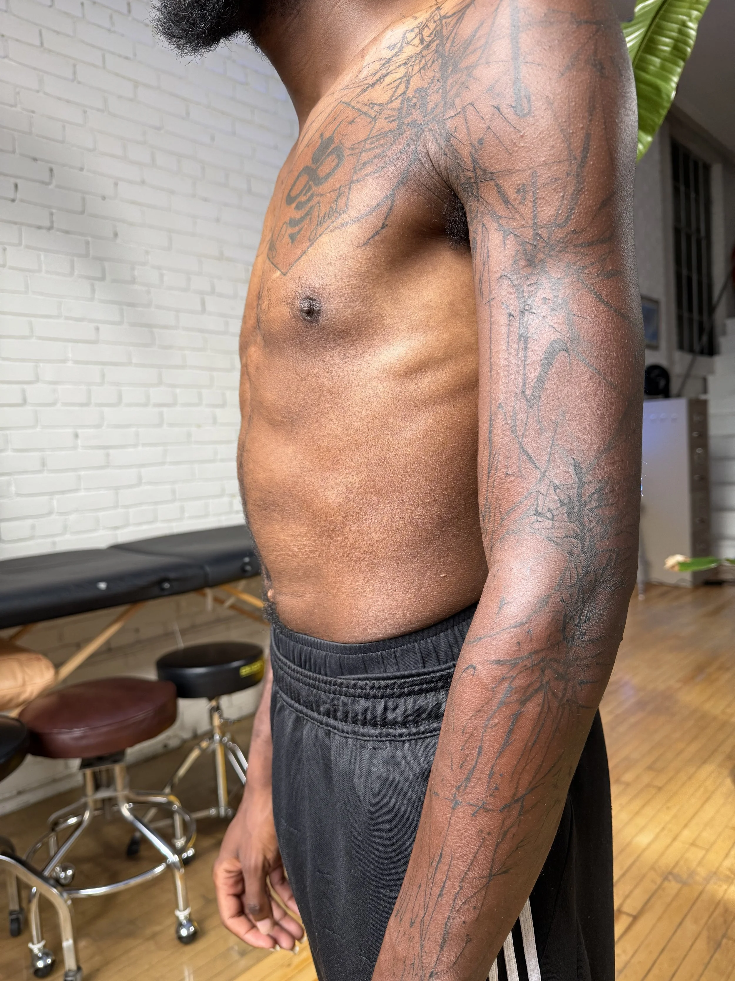 A shirtless person with tattoos on their arm and upper chest, wearing black shorts, standing indoors near a white brick wall and some stools.