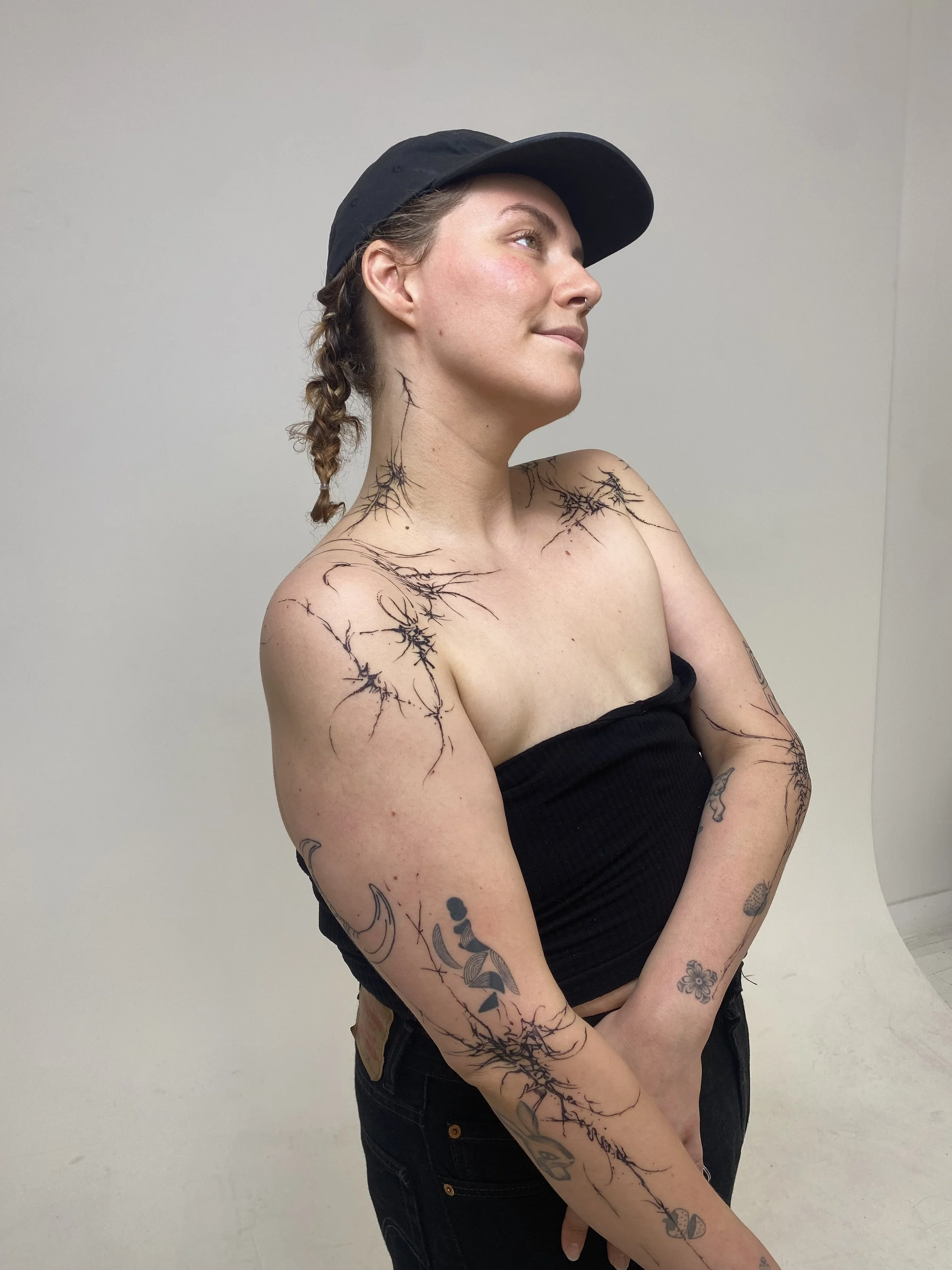 A woman with tattoos on her shoulders, arms, and neck, wearing a black tube top and a black cap, standing against a plain background.
