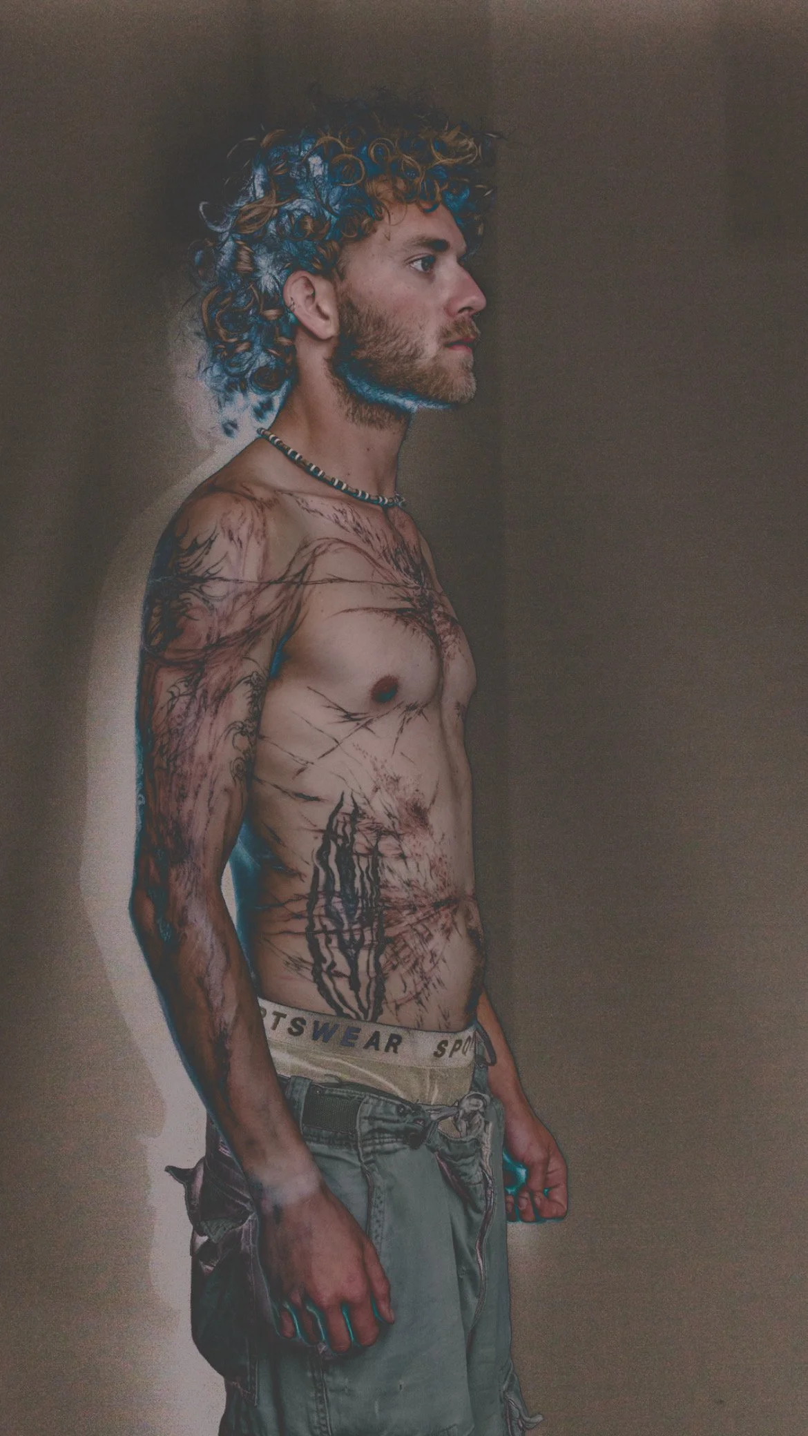 Side view of a shirtless young man with curly hair, numerous abstract flowing tattoos on his arm, torso, and chest, wearing cargo shorts and a necklace, holding a phone in his hand, standing against a plain wall.