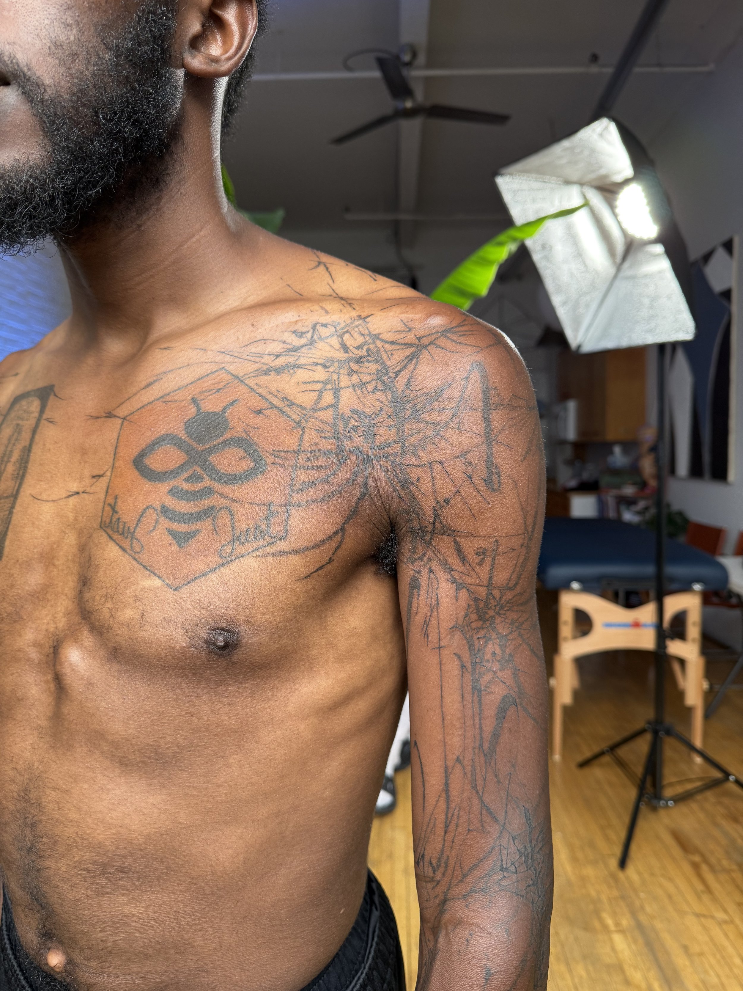 Close-up of a shirtless man with black tattoos on his shoulder and arm, in a photography studio with a softbox light, a height measurement tripod, and a massage table in the background.