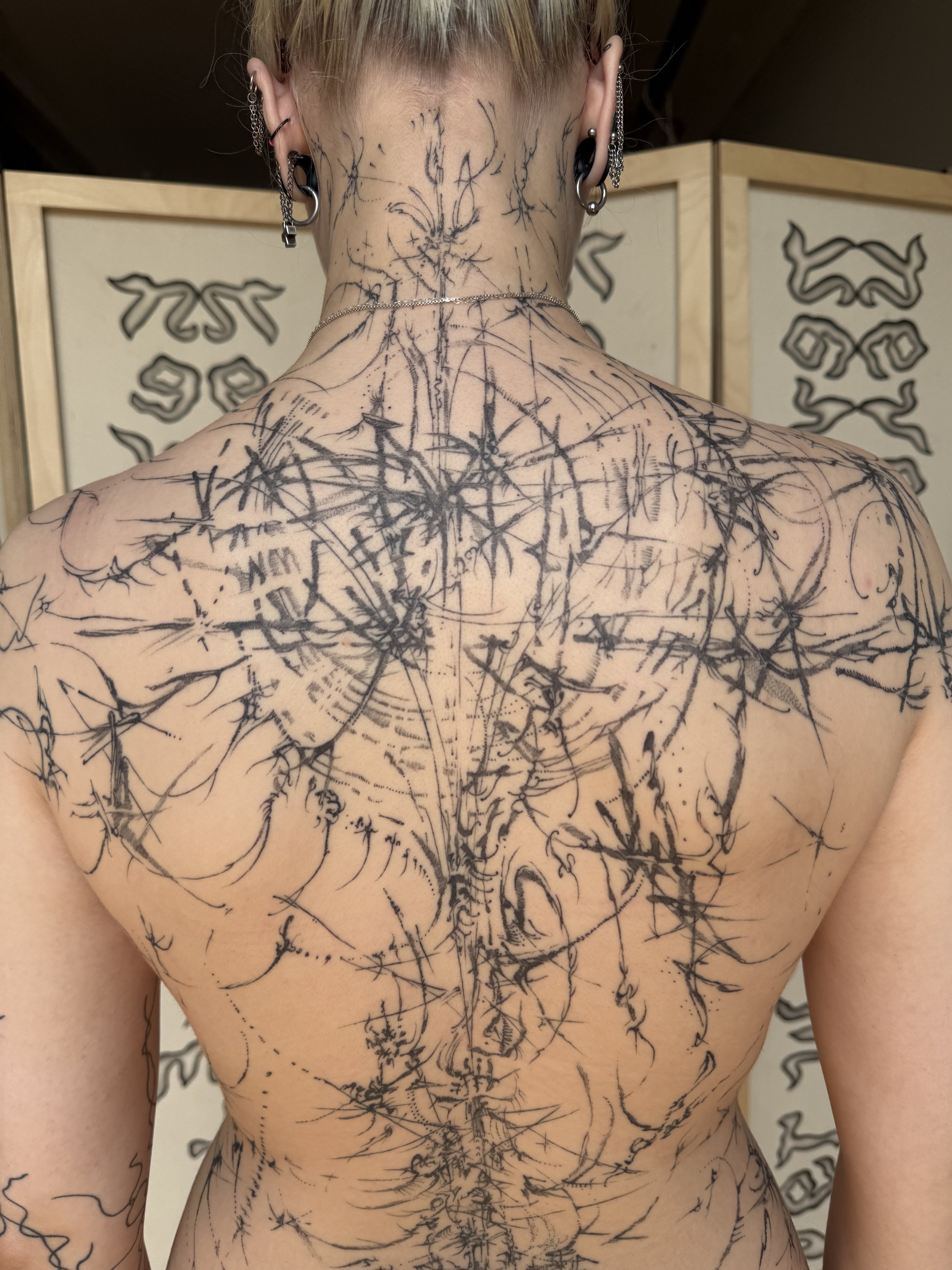 A person with multiple black tattoo outlines on their back and neck, including geometric and abstract designs, in front of a folding screen with similar artistic patterns.