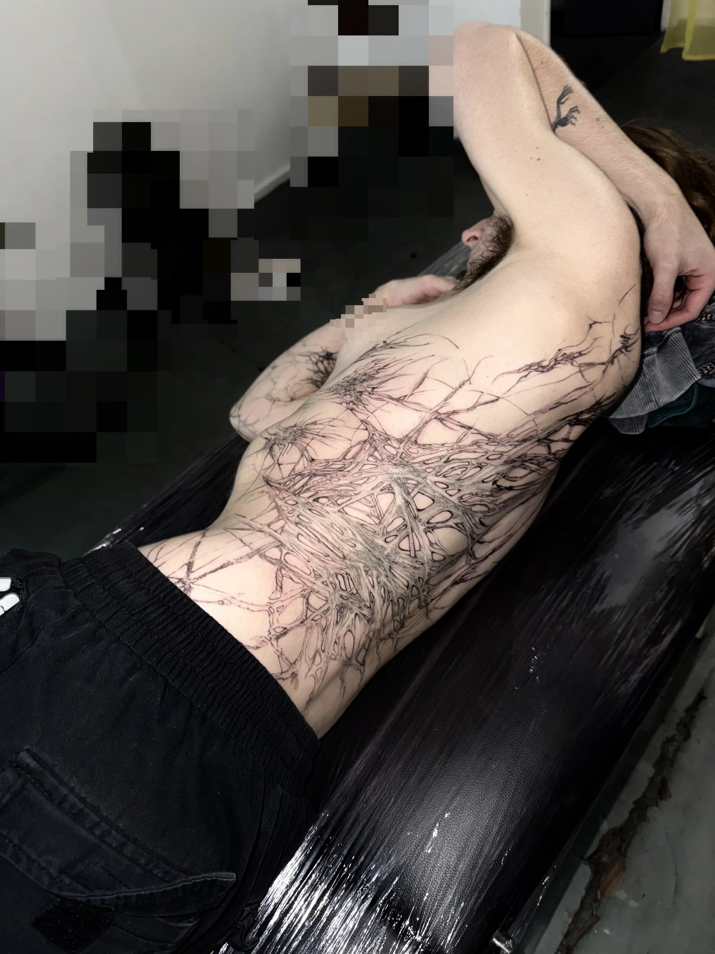A person lying on a tattoo bed with partial upper body and arm tattoos, including a large tree and geometric design, awaiting tattooing.