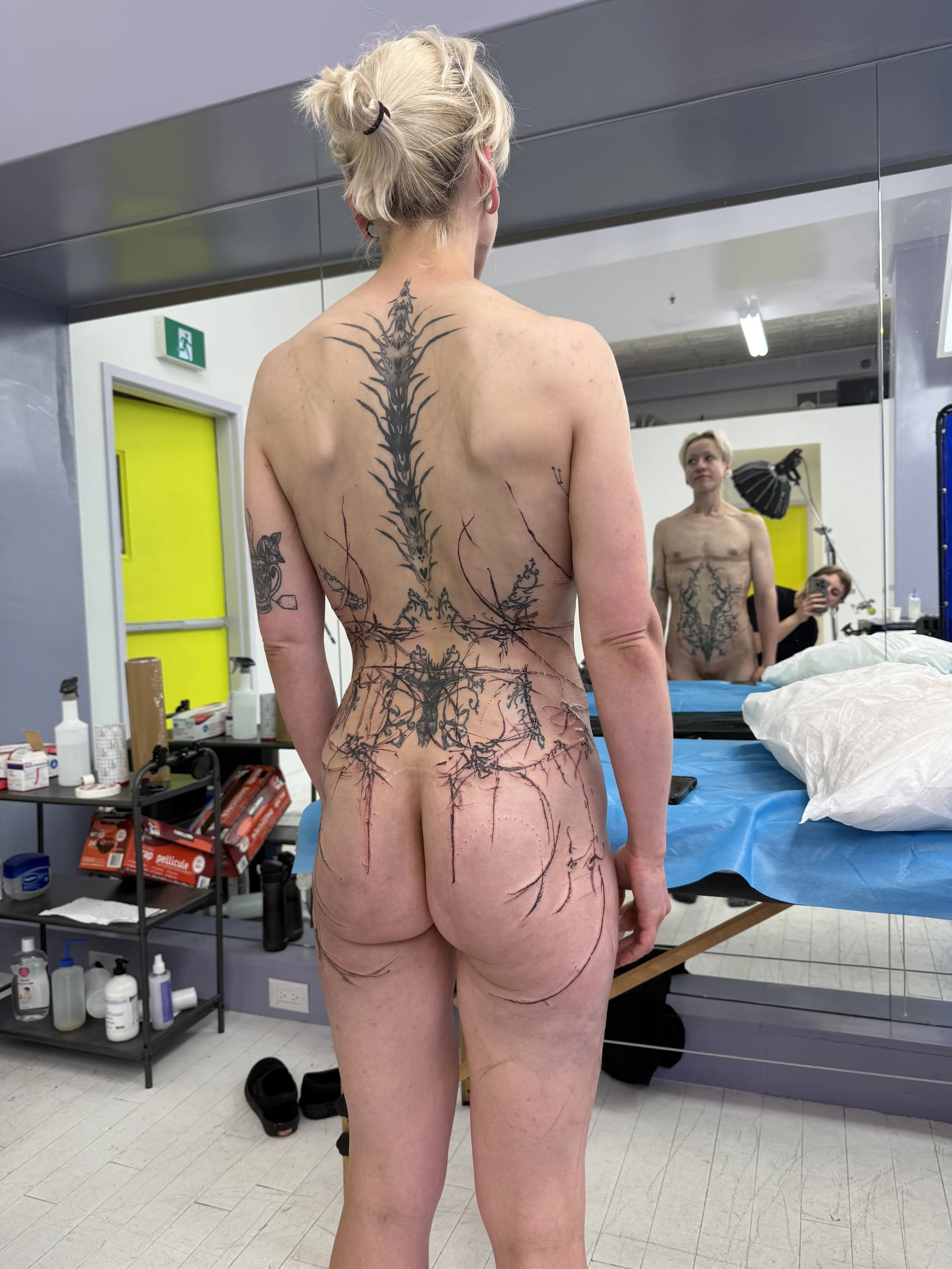 A woman standing in front of a mirror, showing her back with ongoing tattoo work and new inkings on her shoulder, back, and buttocks. The setting appears to be a tattoo studio with various supplies visible on shelves and a blue cover on the table.