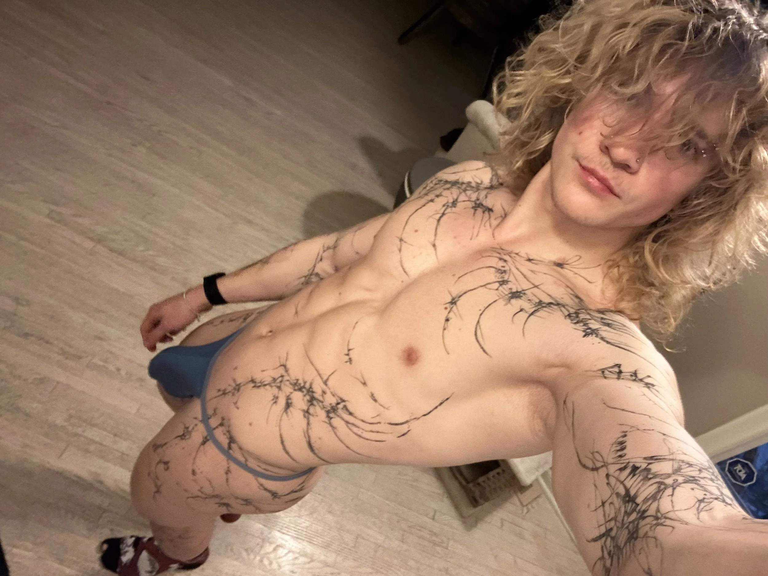 A shirtless young man with curly blonde hair taking a selfie in a room with wooden flooring. He is wearing a blue thong, a black wristband, and socks. The man has tattooed arms, shoulders, chest, and torso with black, thorn-like designs.