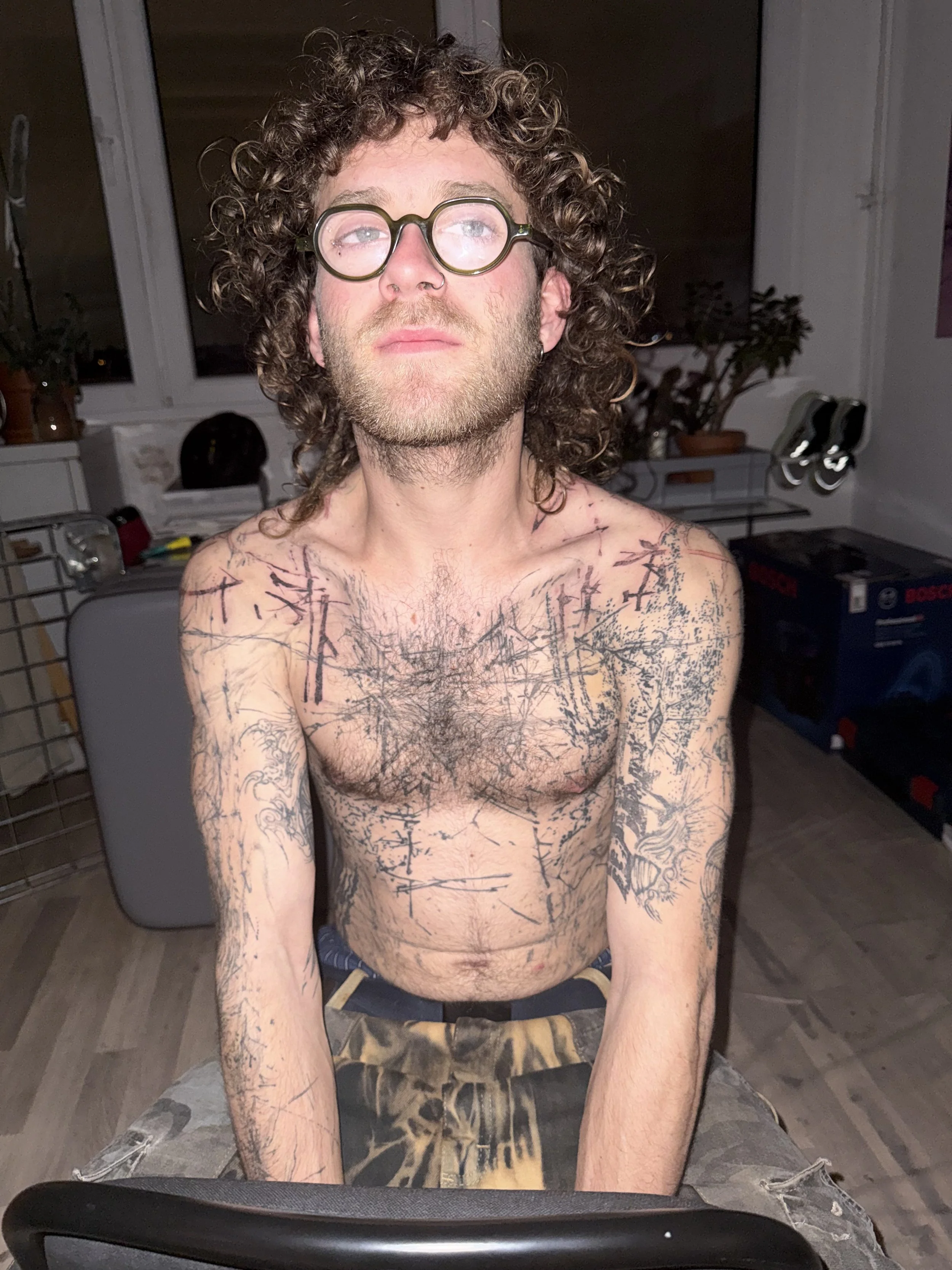 A shirtless man with curly hair, glasses, and tattoos on his chest and arms, sitting indoors with a neutral expression.