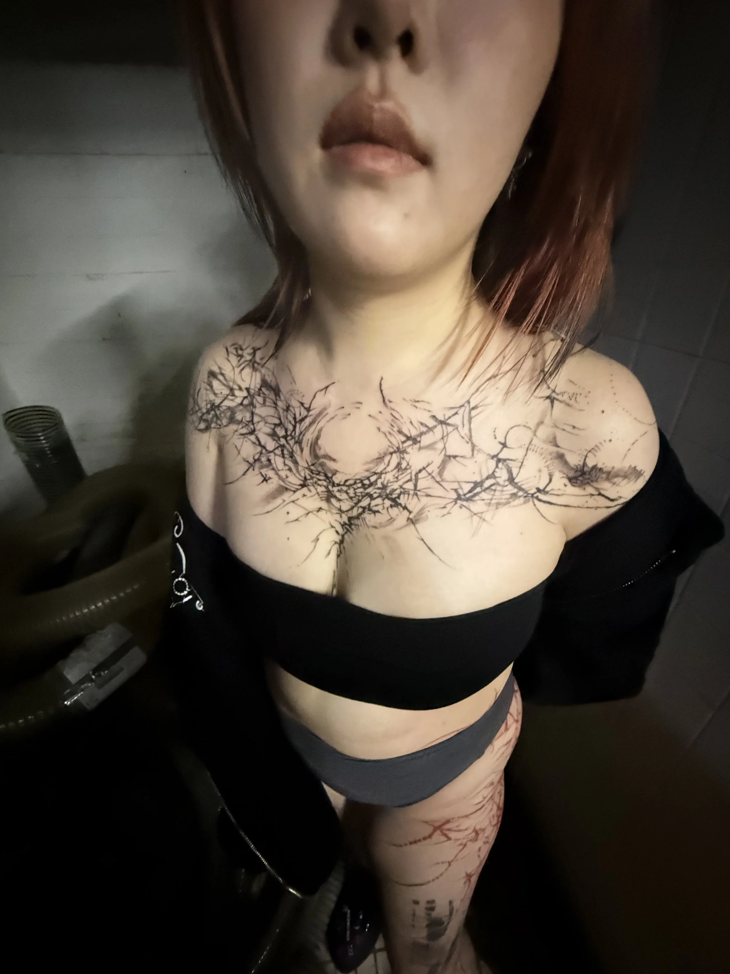 A woman with shoulder-length red hair and tattoos on her chest and right thigh is taking a selfie from a high angle, showing her face, chest, and one leg. The background appears to be a tiled room, possibly a bathroom.