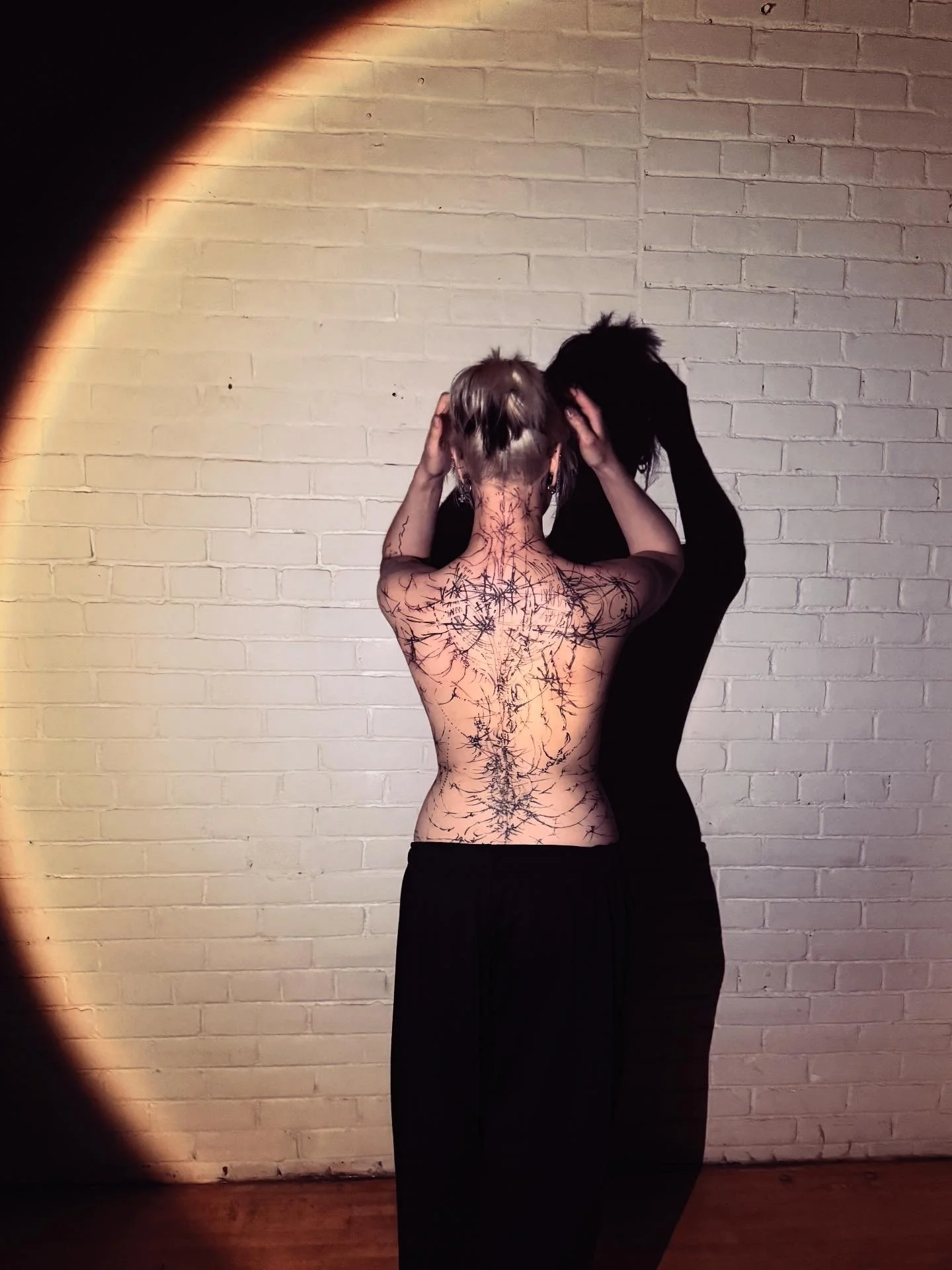 A person with short, light-colored hair facing a light brick wall, holding their head with both hands, with a black tattoo design covering their back and extending to their arms. The person's shadow cast on the wall is visible, and they are wearing b