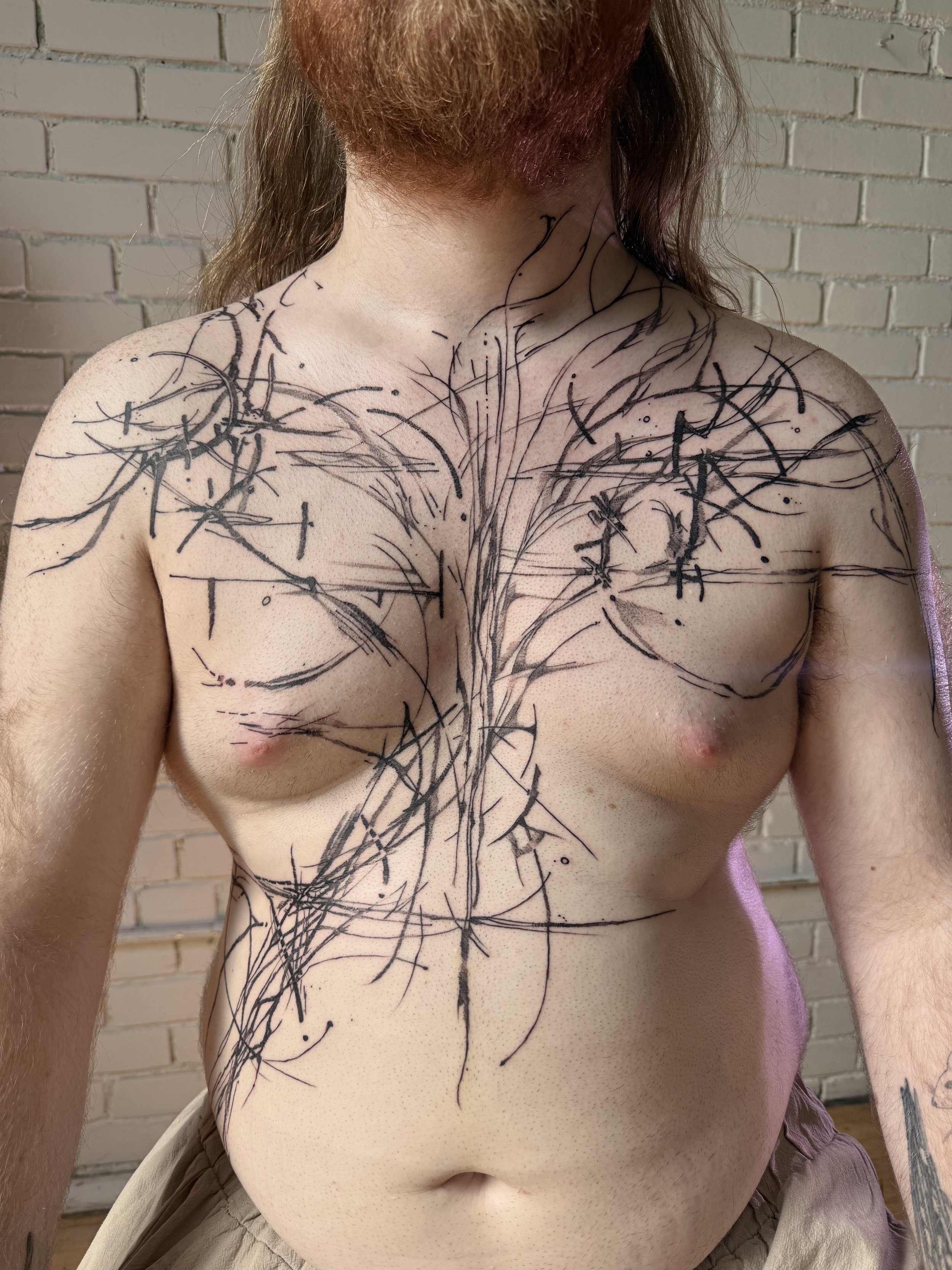 A shirtless man with a beard and long hair has abstract black tattoos drawn all over his chest, shoulders, and arms. The tattoos consist of lines, circles, and other geometric shapes, creating a layered, intricate design.