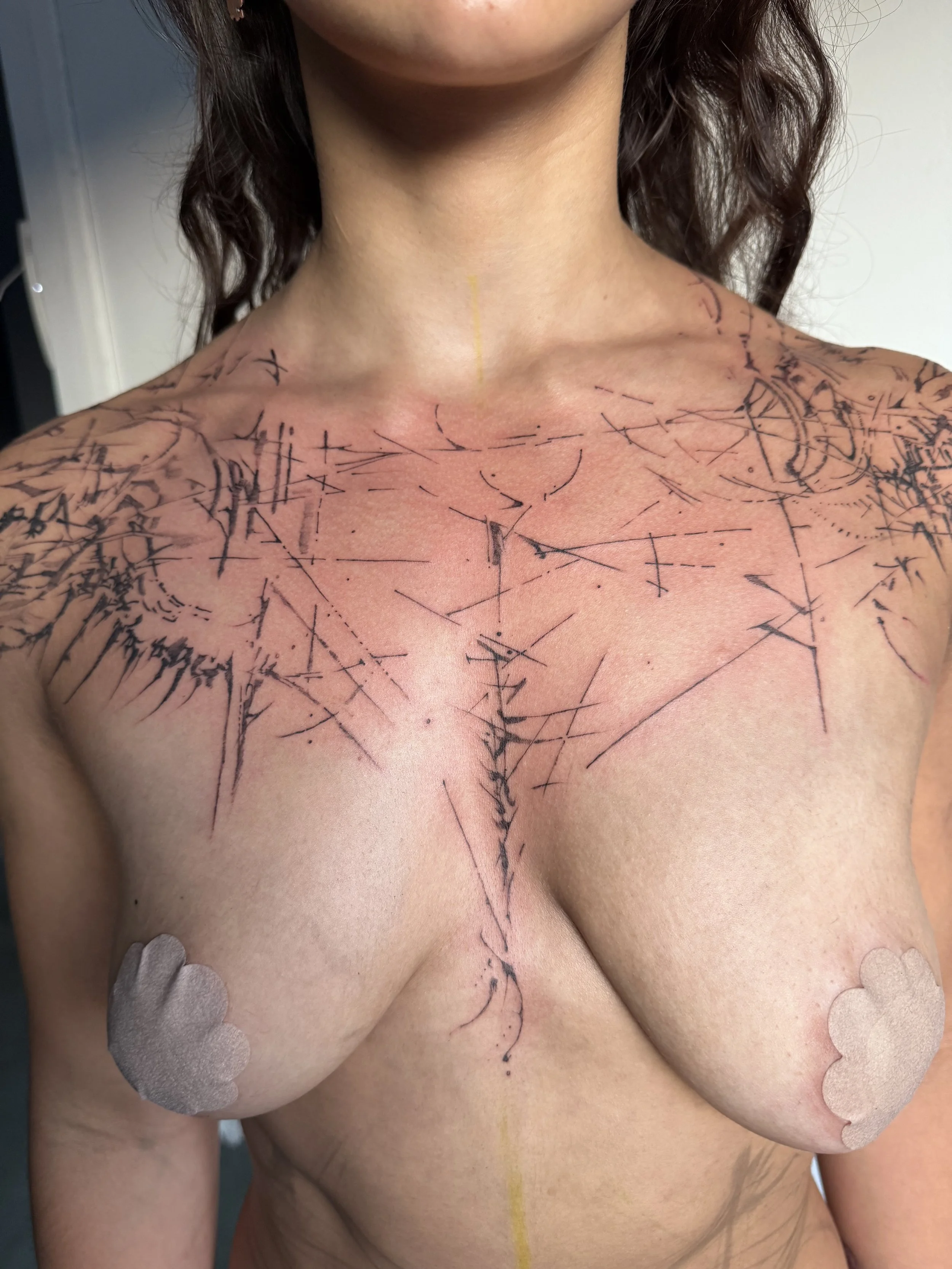 A woman with dark, curly hair and light skin has a recent tattoo on her shoulder and chest, consisting of black abstract lines and shapes that resemble a shattered glass or spider web pattern and conform to the lines of her body.