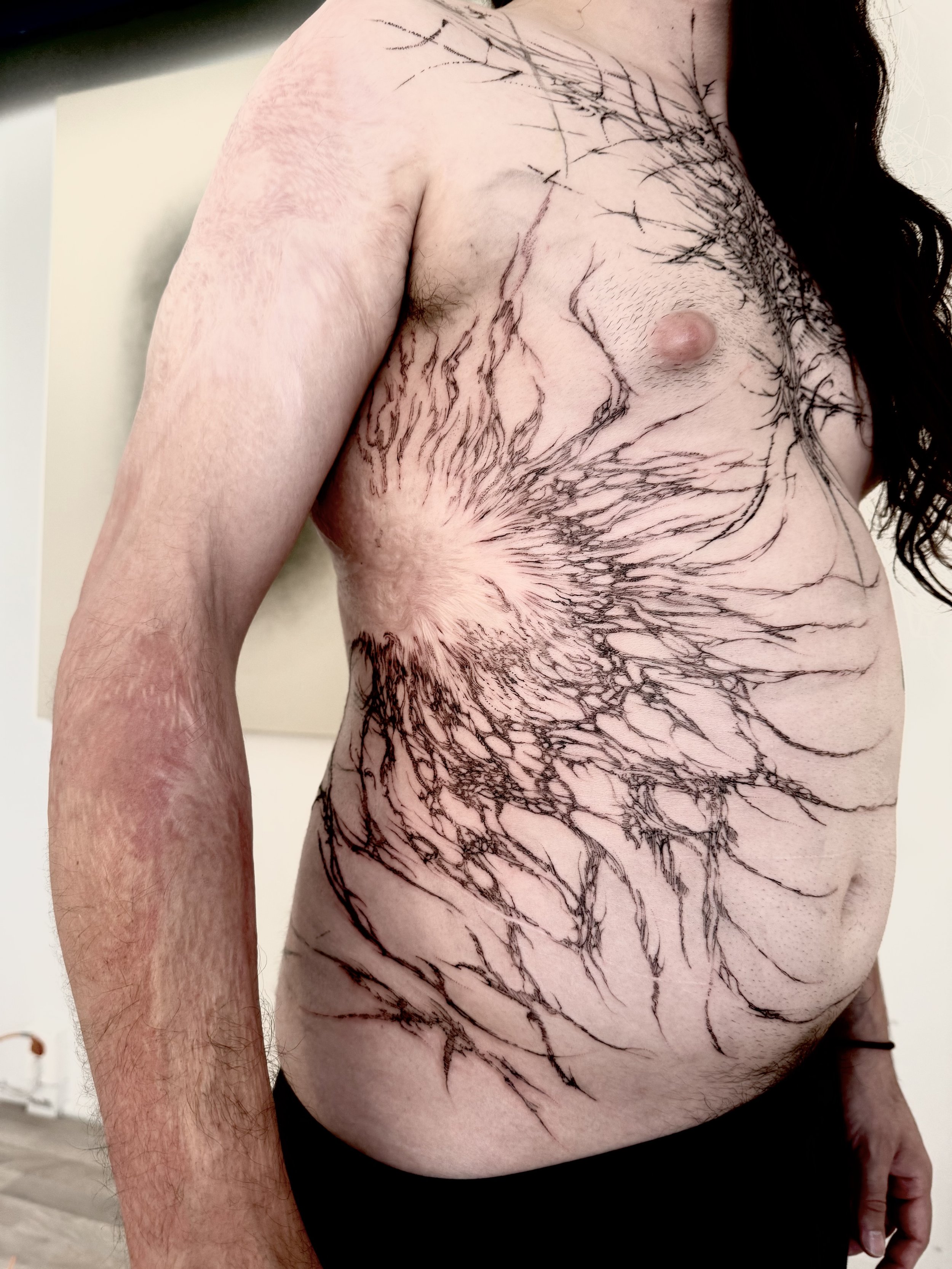 Close-up of a person's chest, arm, and side, with intricate black ink tattoo resembling tree branches or roots.