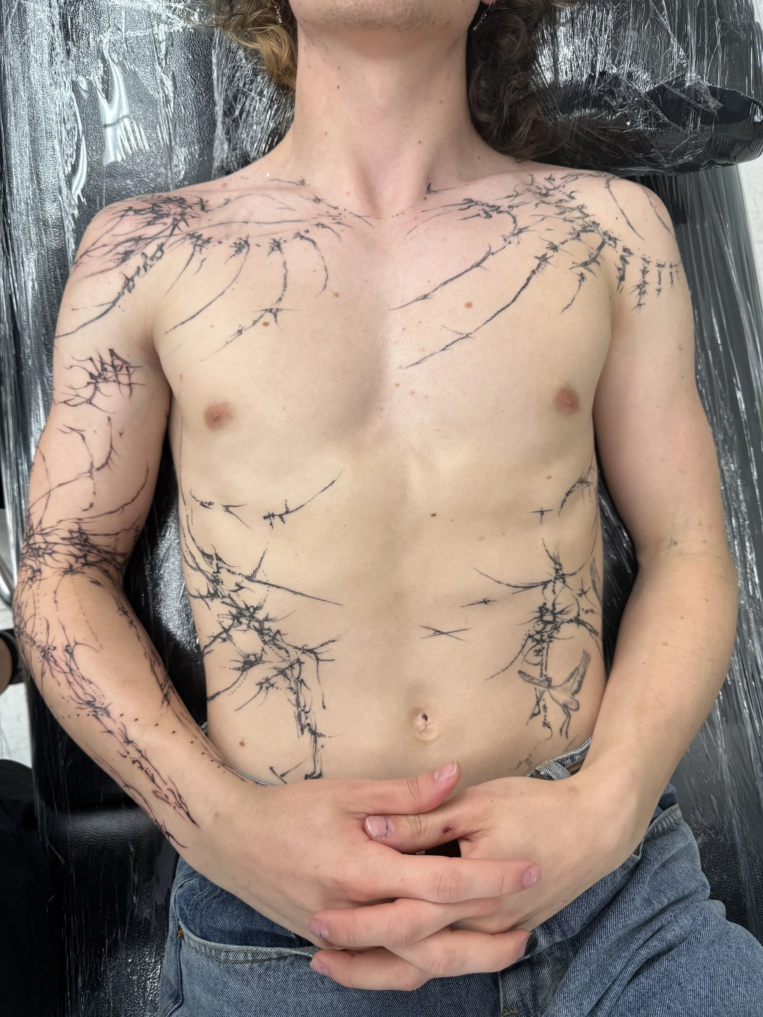 A shirtless person with multiple black line tattoos that resemble lightning or tree branches across their chest, shoulders, and arms, lying on a black surface covered with plastic.