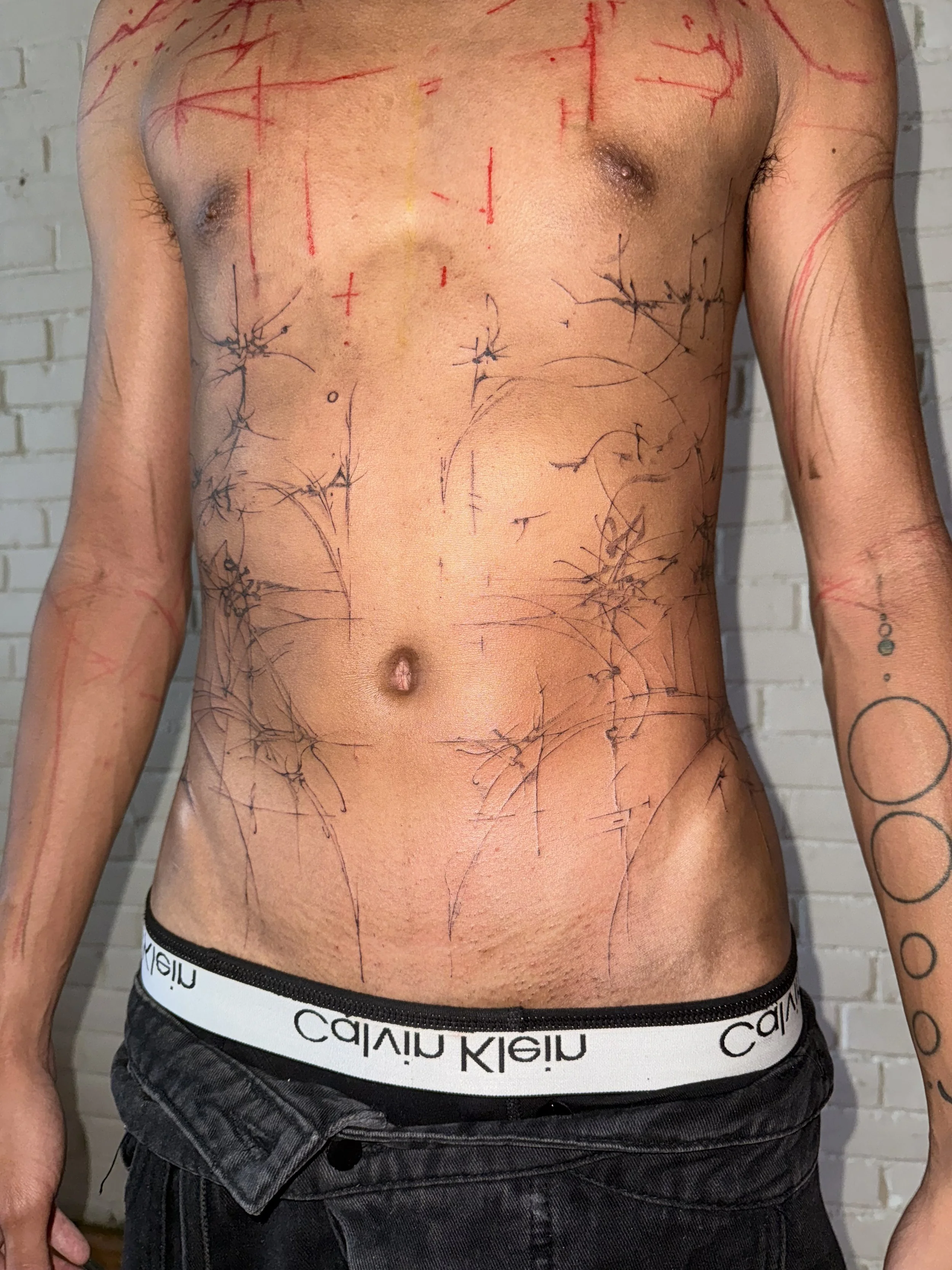 A shirtless person with tattoos and red and black ink sketches on their body, standing against a white brick wall.