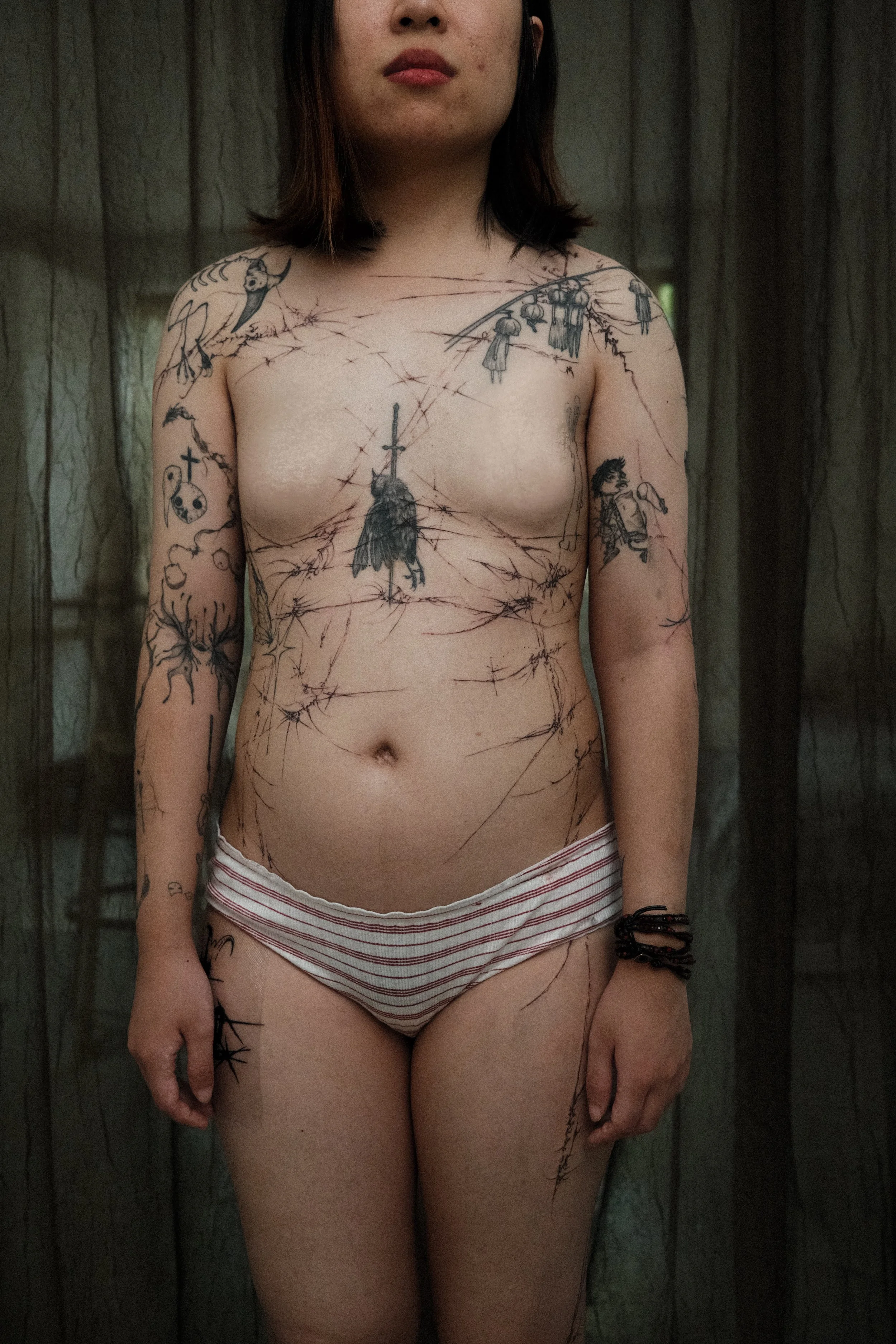 A woman with tattoos covering her upper body, standing with a neutral expression in front of dark curtains.