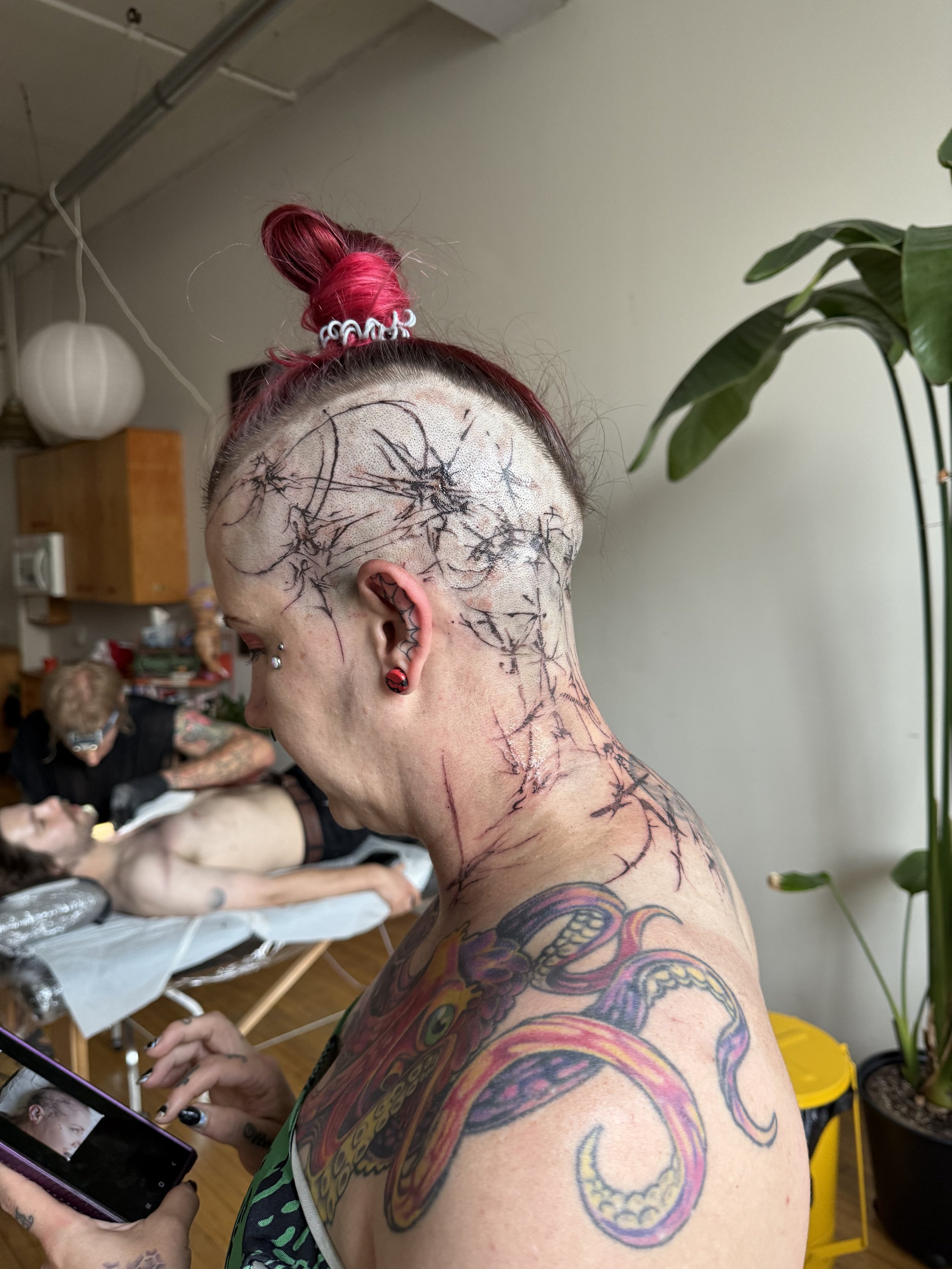 A woman with tattoos and a shaved head, with black ink spider web designs on her head and neck, taking a selfie in a tattoo studio with a person lying on a tattoo bed and a tattoo artist working on them in the background.