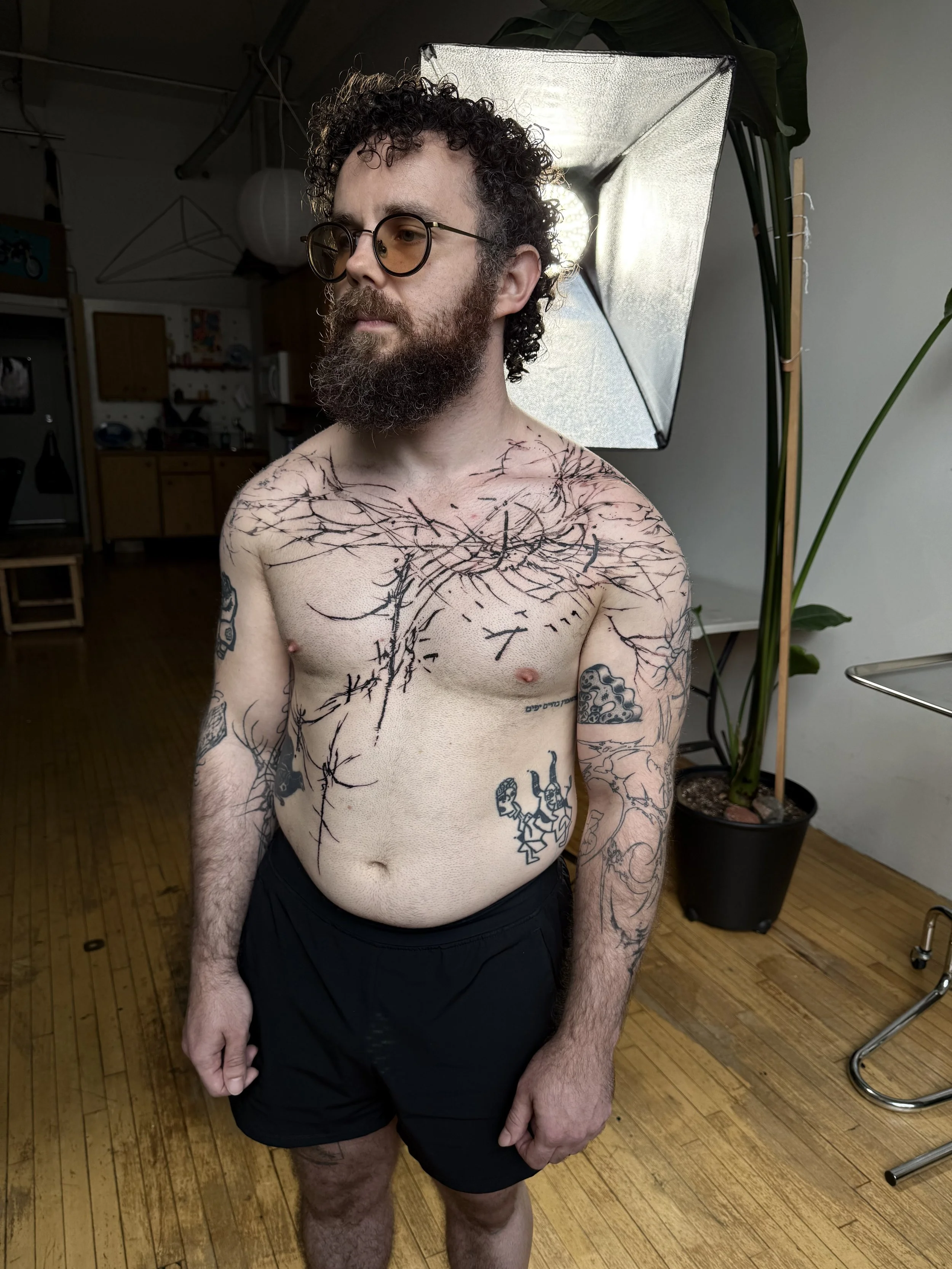 A shirtless man with curly hair, glasses, and a beard, standing indoors in front of a photography setup with a softbox light. He has multiple tattoos on his chest, arms, and torso, including a large tree with branches across his chest and various sma