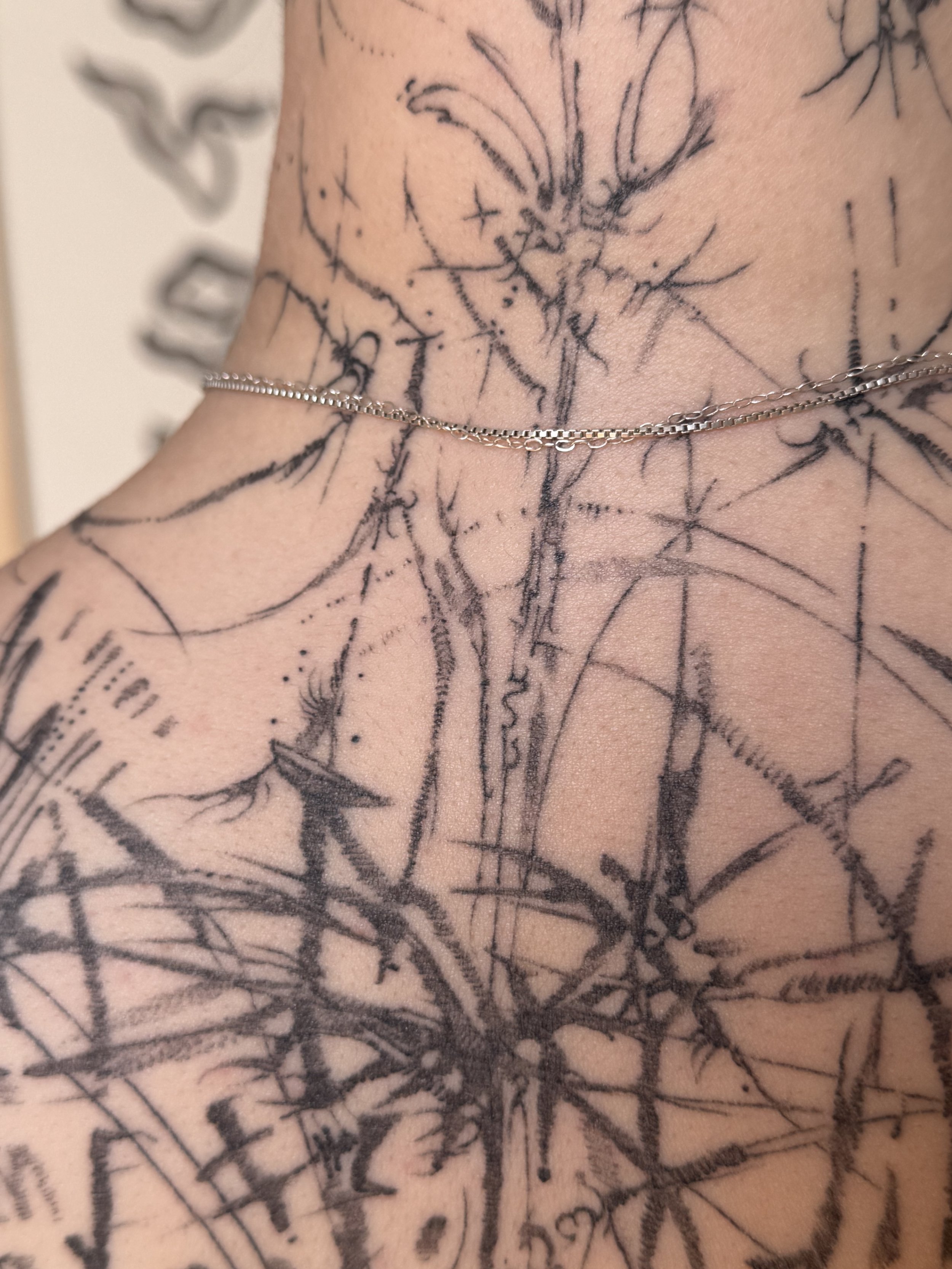 Tattooed person's neck and shoulder with abstract black ink designs and a silver chain necklace.