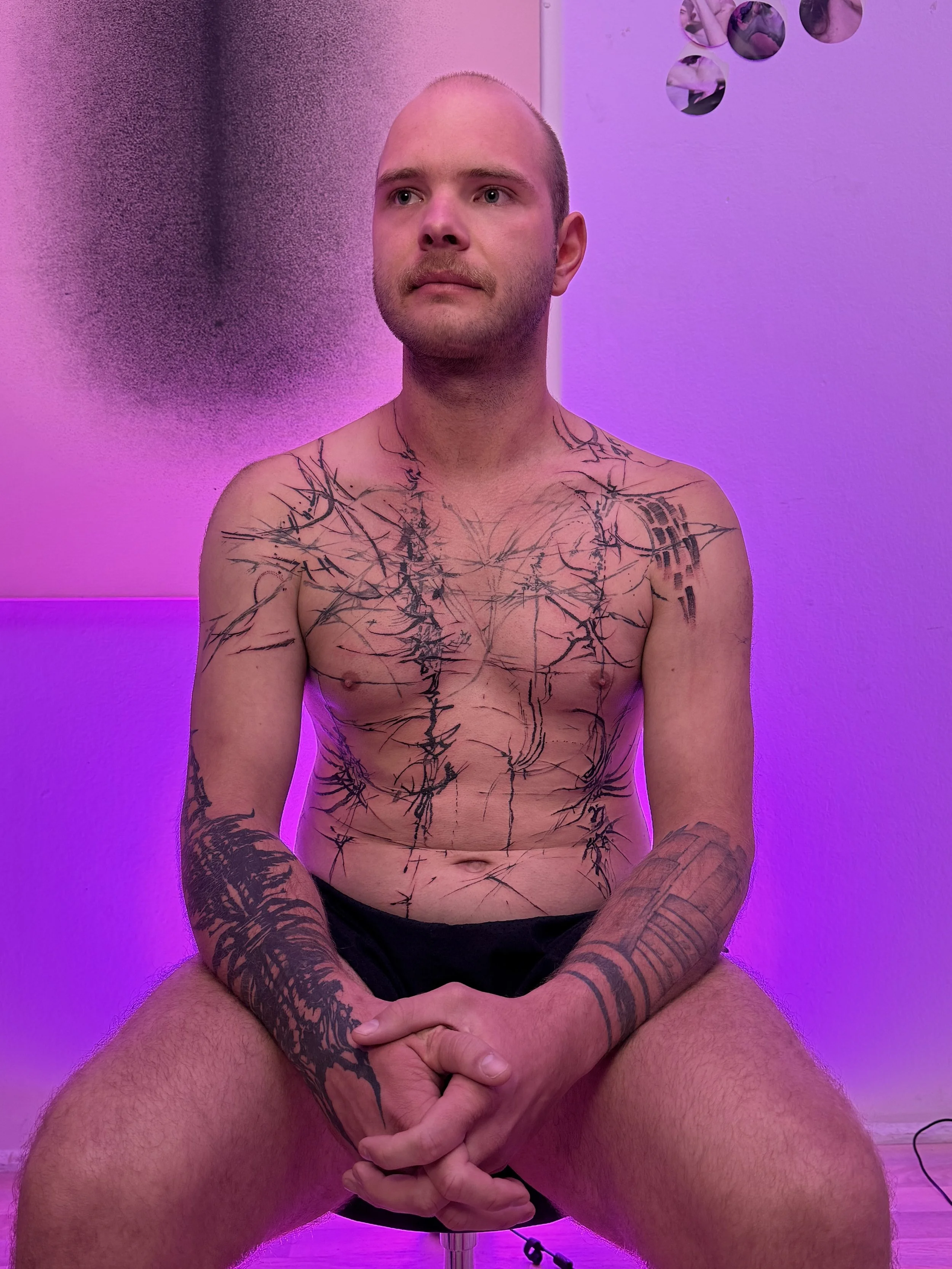 A shirtless man with numerous tattoos, sitting in a chair with hands clasped. The background has pink and purple lighting, and there is wall art behind him.
