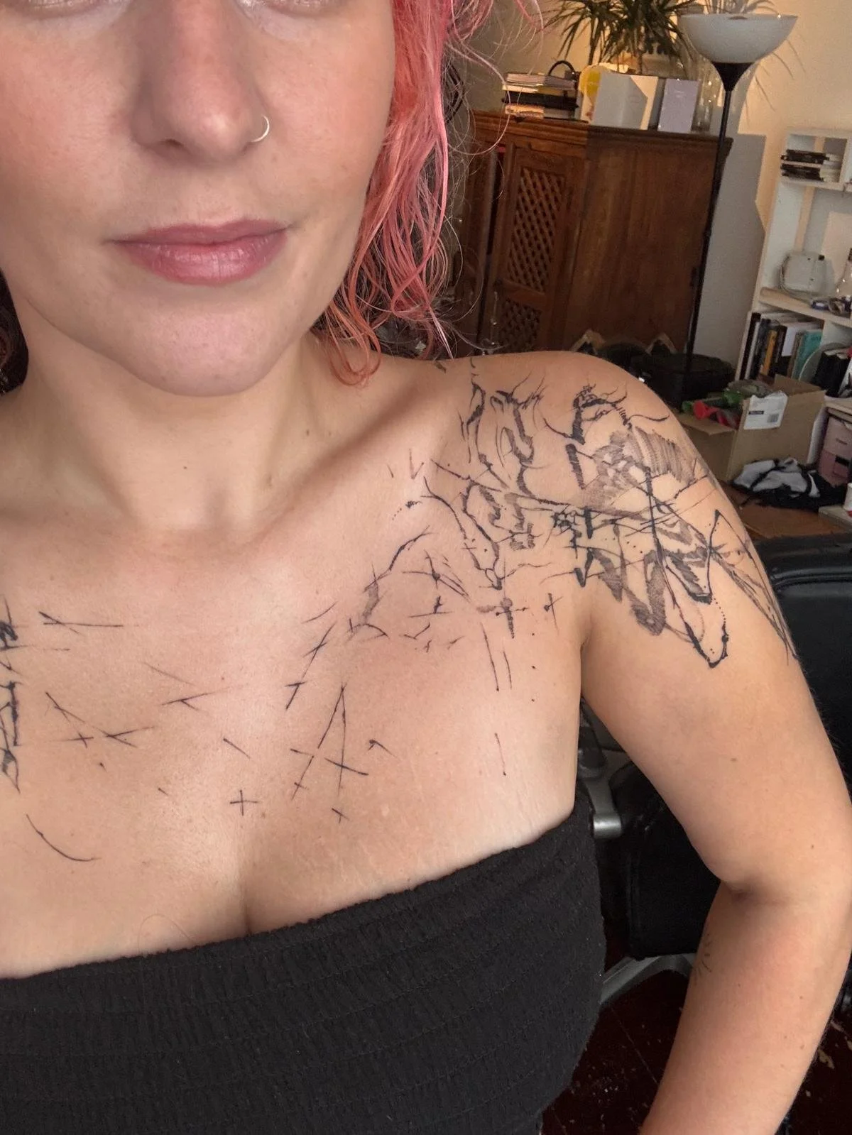 A woman with pink curly hair and a nose ring smiling in a room with books and furniture, showing a large, abstract black tattoo on her chest and shoulder.