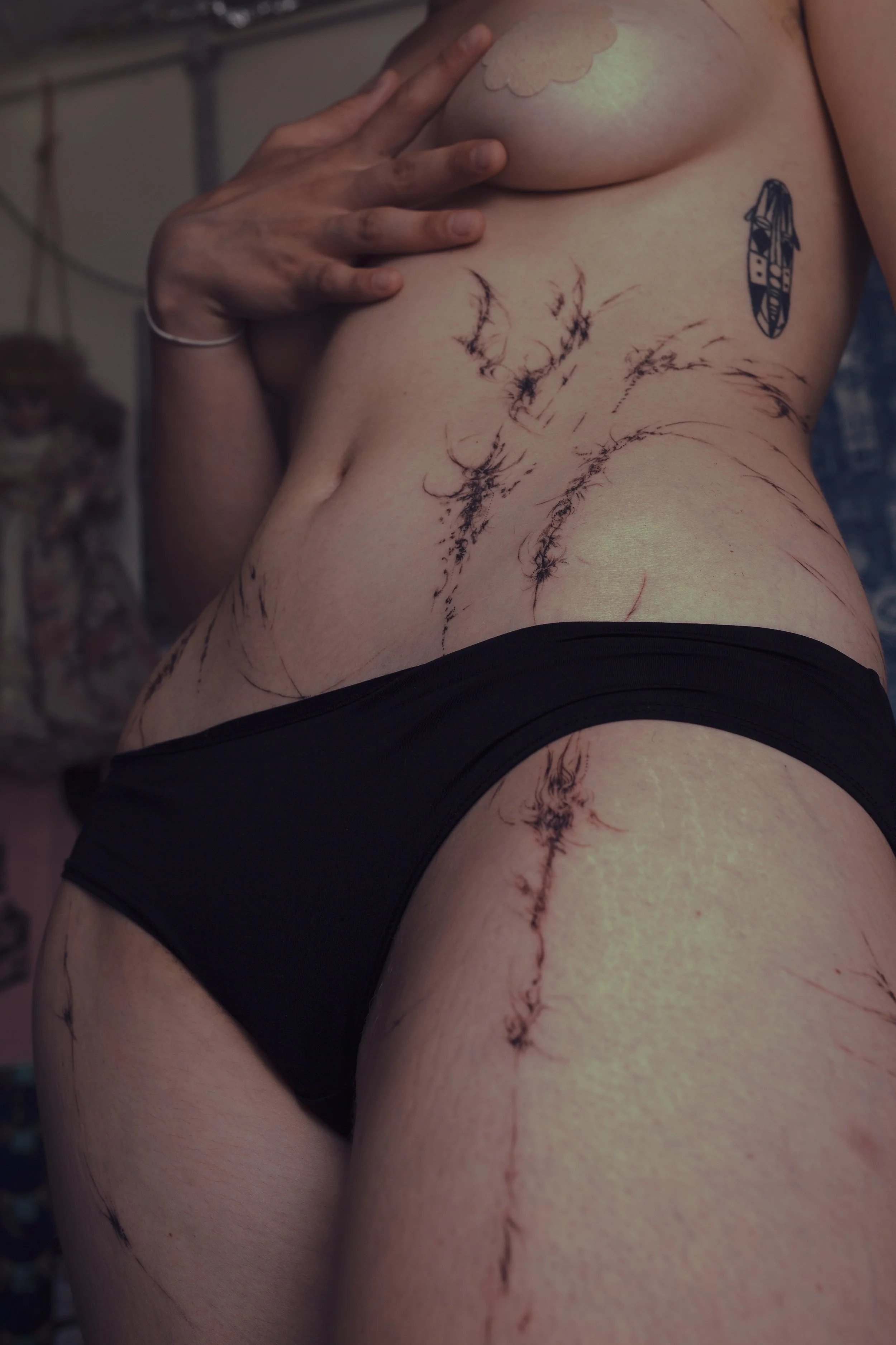 A woman with multiple delicate and fine line tattoos on her torso, side, and hip, partially covering her breast with her hand. She has abstract tattoo on her upper chest and is wearing black underwear.