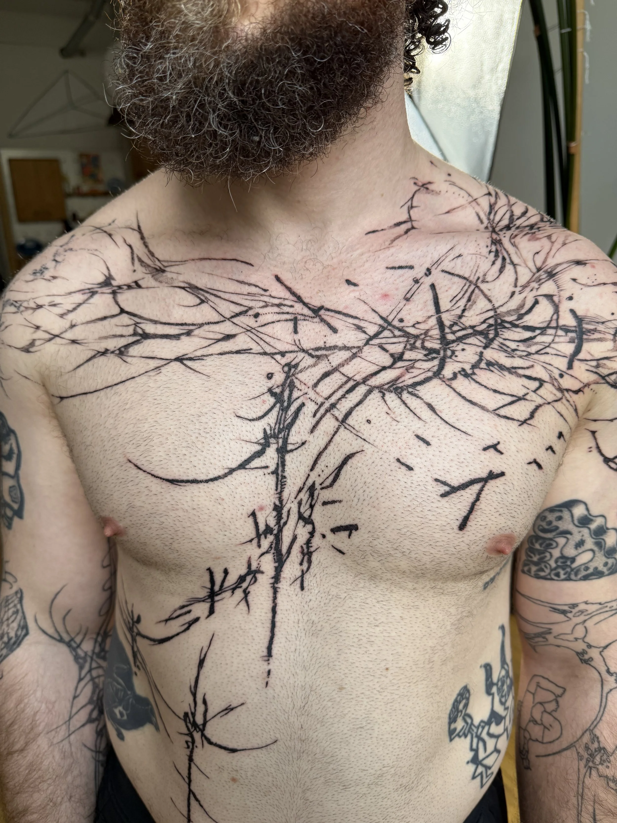 A shirtless person with a beard and curly hair displaying fresh black ink tattoos that resemble tree branches or abstract lines across their chest and shoulders, with additional tattoos on their arms.