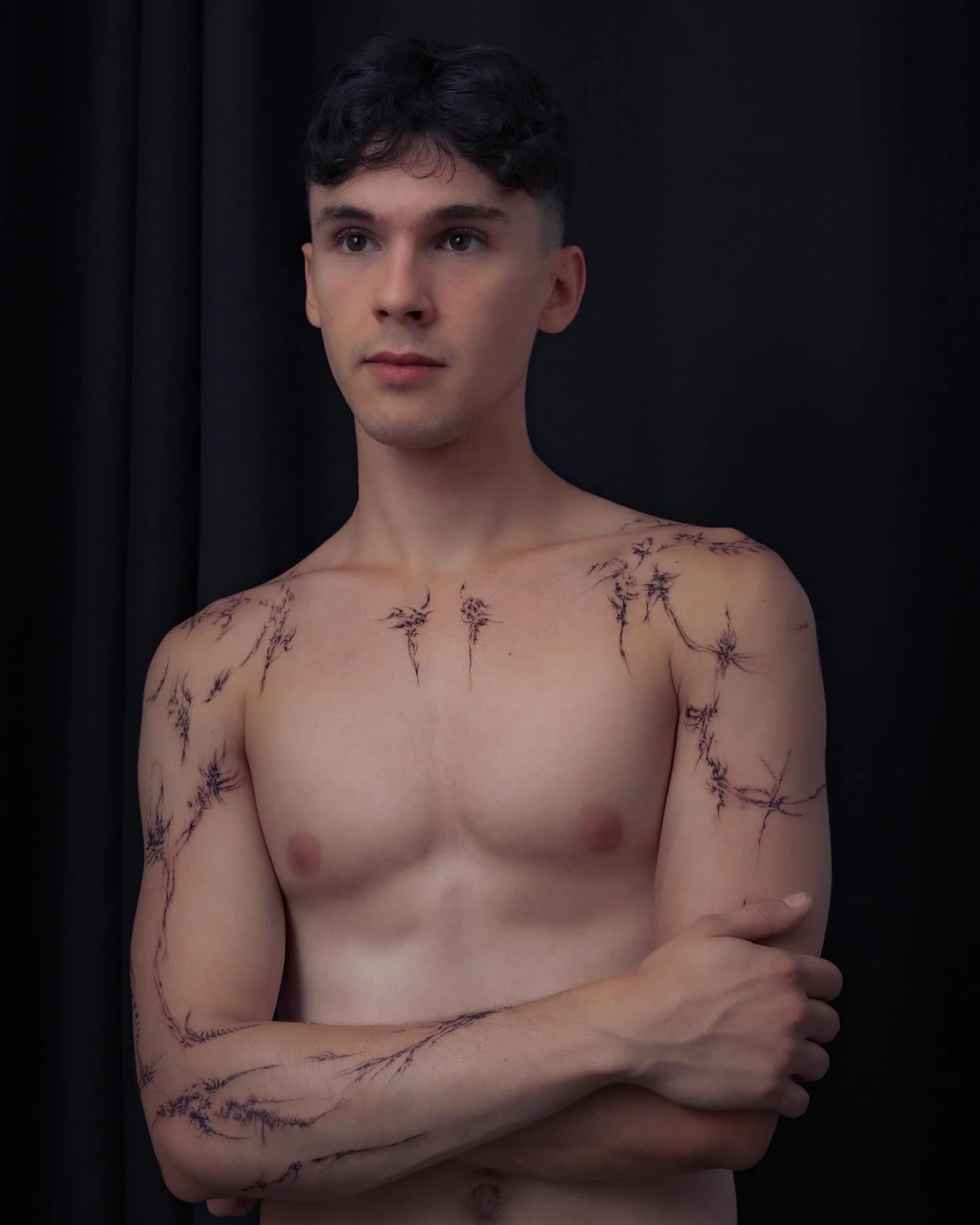 A shirtless young man with dark hair and light skin stands against a black background, with tattoos resembling lightning bolts or jagged lines on his chest and arms.