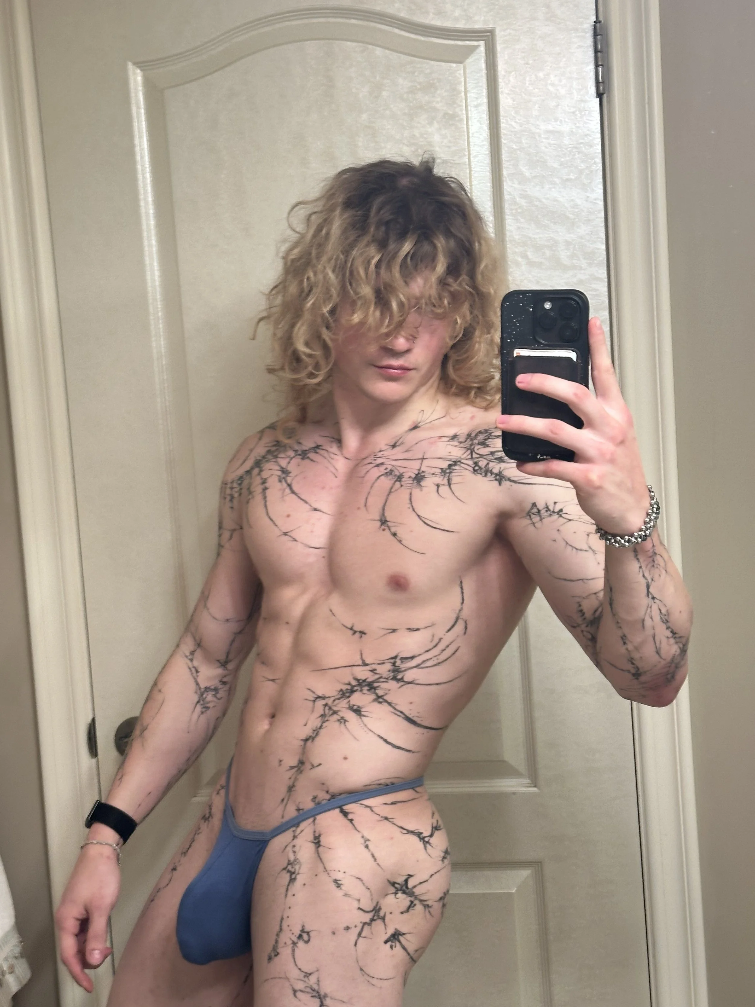 A shirtless man with curly hair taking a selfie in a mirror, wearing blue underwear and a black wristband, with black ink tattoos resembling branches across his chest, arms, and torso.