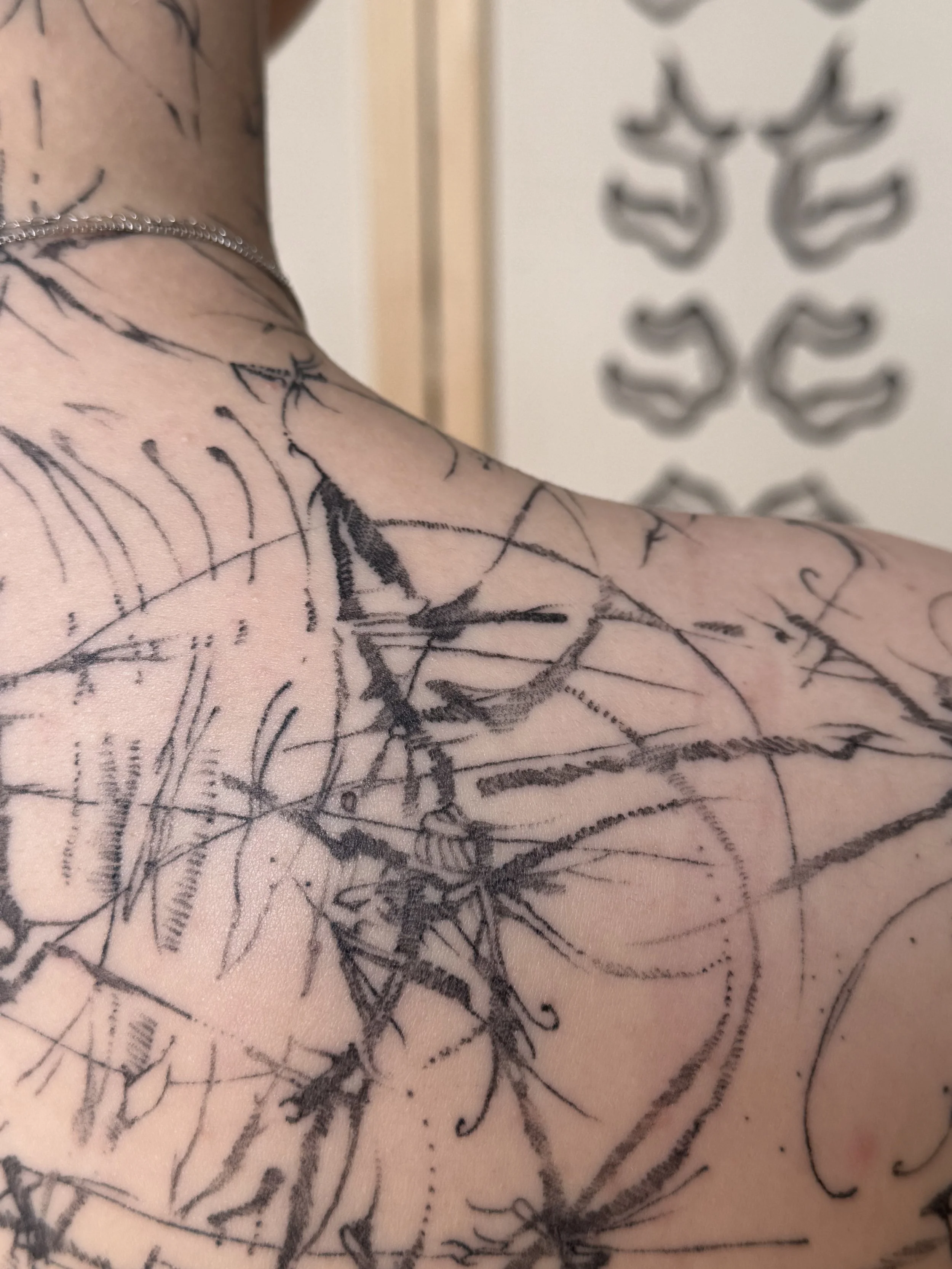Close-up of a person's shoulder and upper chest with multiple black ink tattoos, including abstract lines and shapes, with part of a necklace visible.