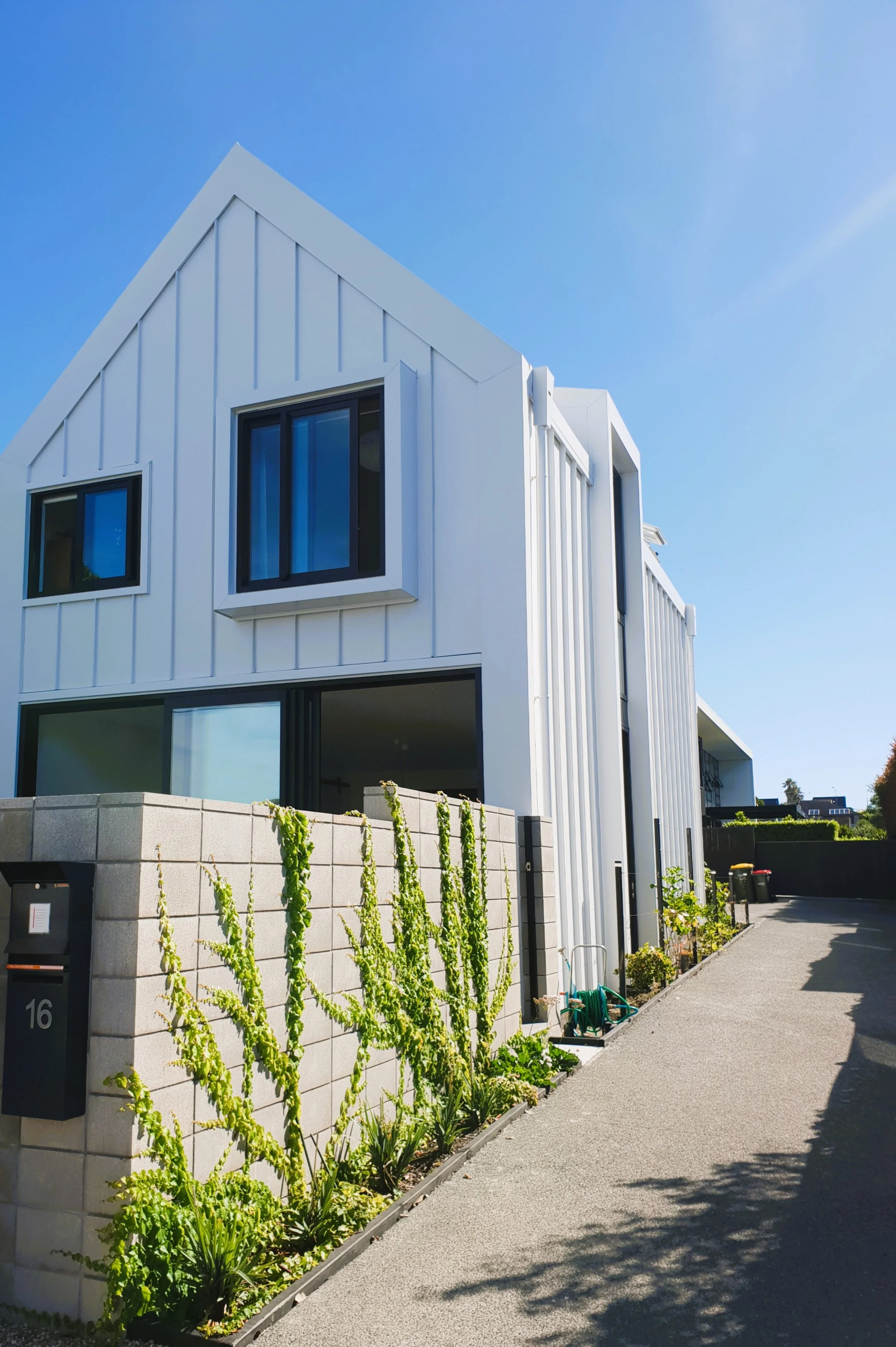 Modern white two-story house with large windows, a concrete block fence, and a sidewalk with plants and garden tools under a clear blue sky.