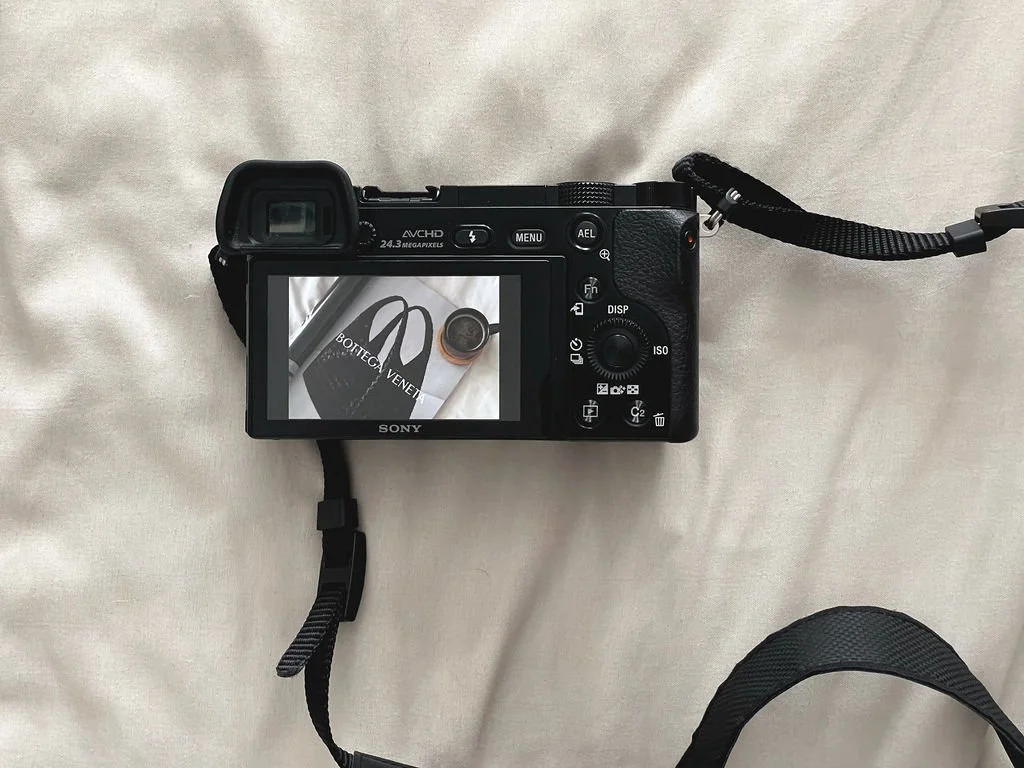 A Sony digital camera on a light-colored fabric surface, with a strap attached. The camera's LCD screen displays a photo of a Bottega Veneta item, including a black bag and a round jar.
