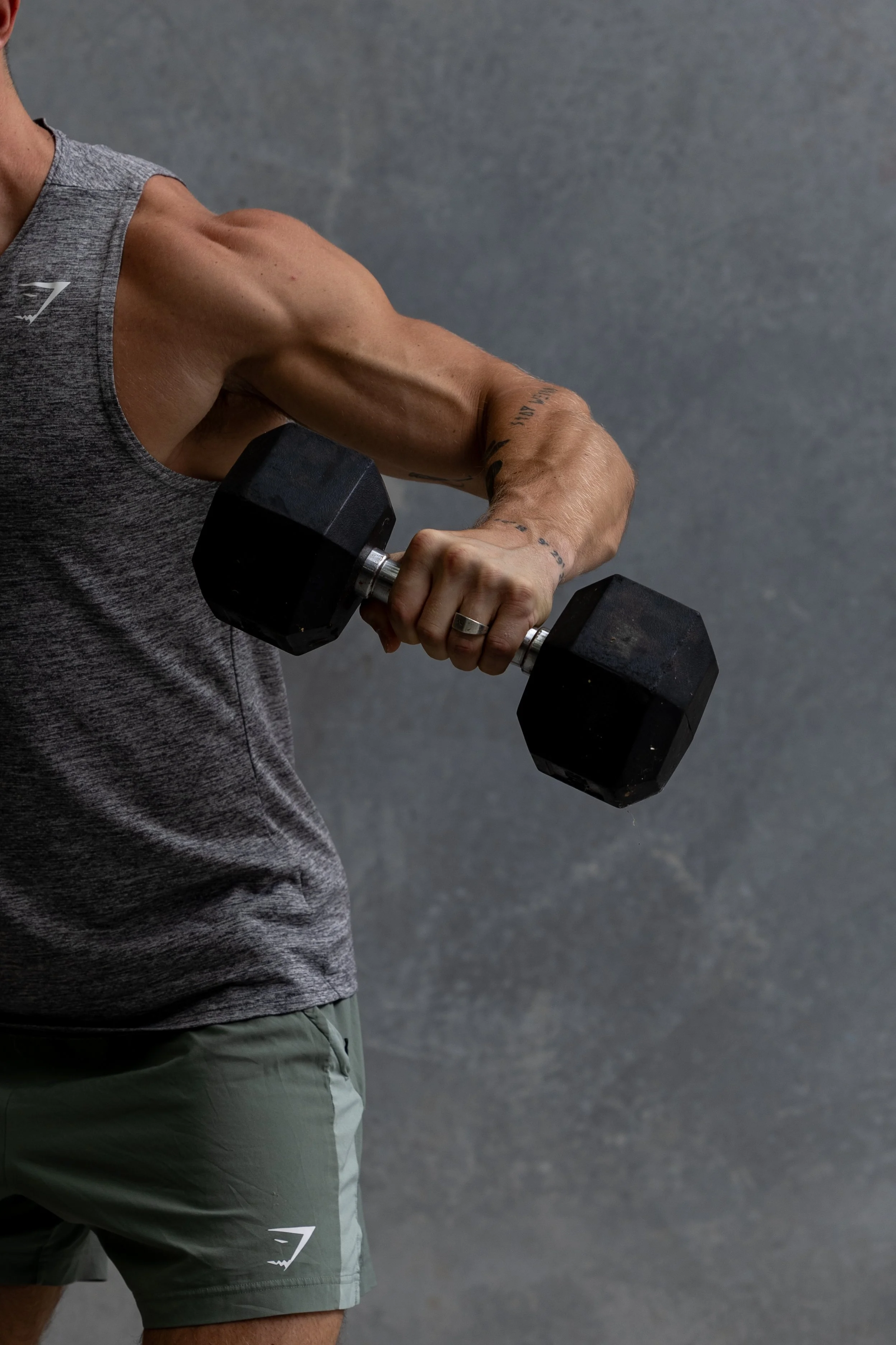 Muscular man in grey shirt and green shorts lifting a large dumbbell, representing wellness content for a personal trainer with virtual assistant support for digital product launches, content marketing, SEO, and admin services
