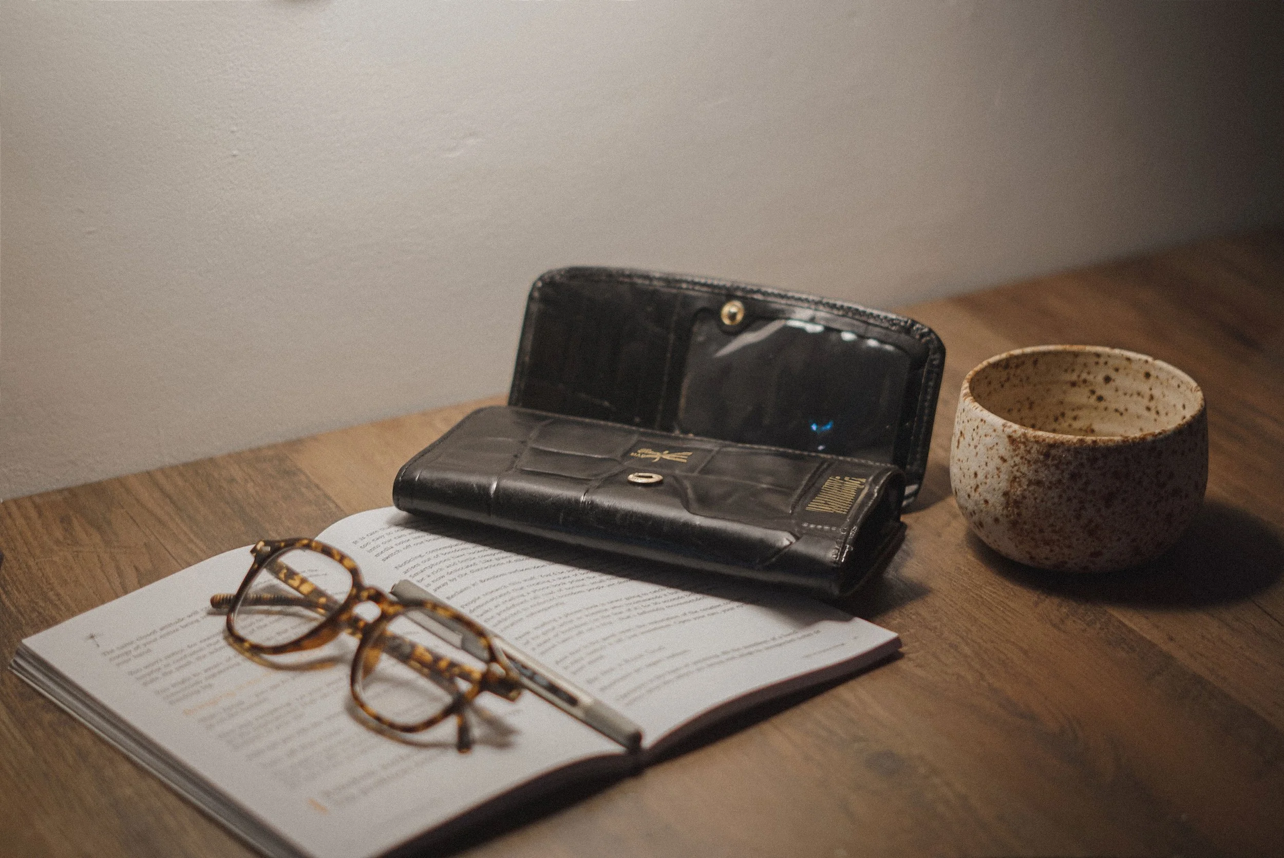 Open book with reading glasses, a black wallet, and a ceramic mug on a wooden table next to a textured wall.