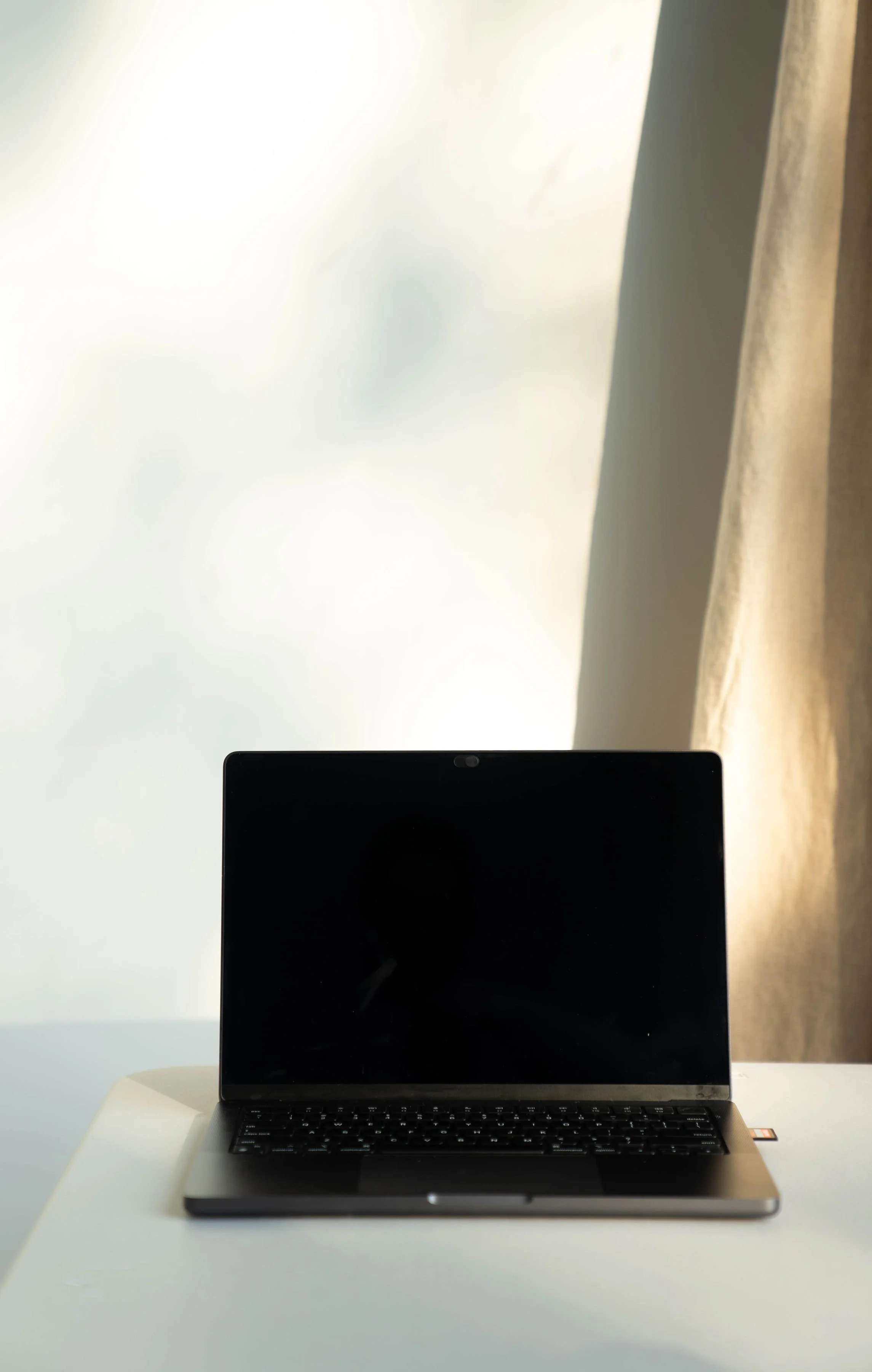 A laptop computer with a black screen on a white table near a window with beige curtains.