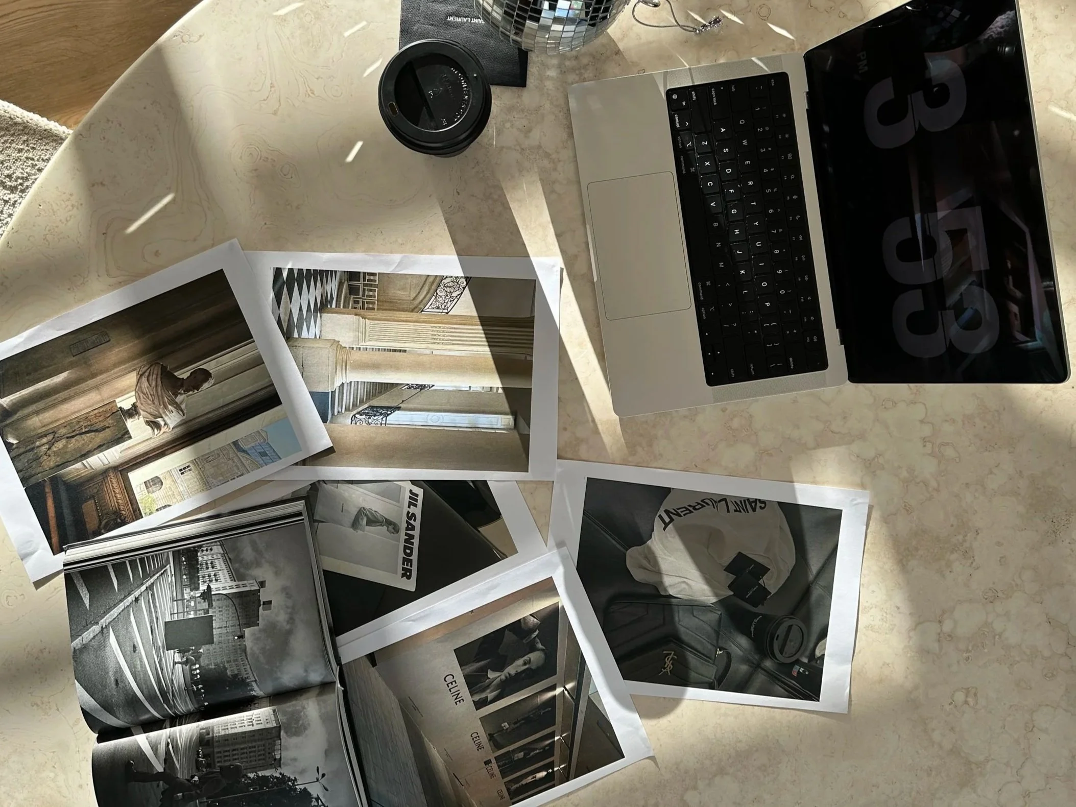 A table with a laptop, a coffee cup, printed photographs, and a magazine featuring black-and-white city photos.