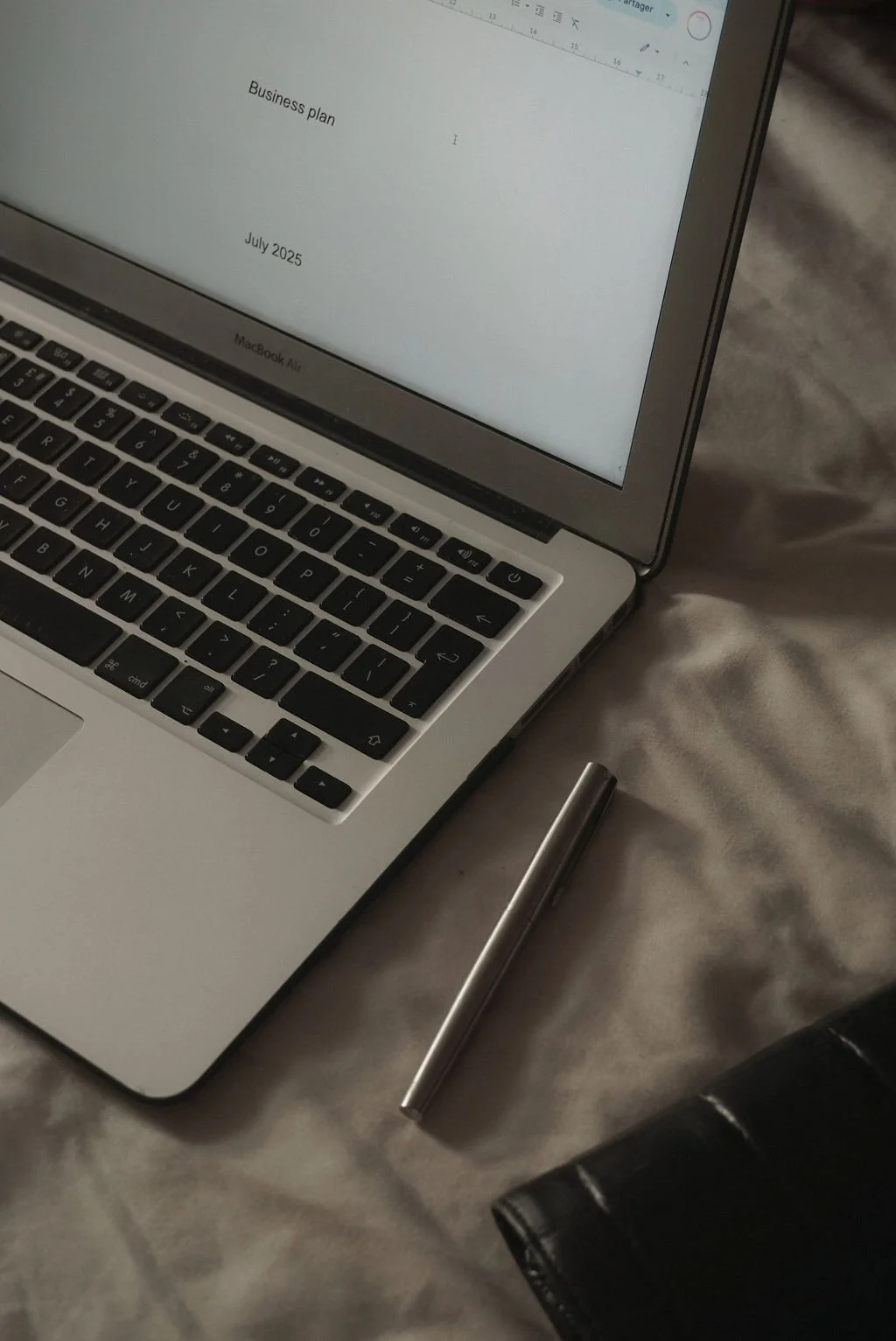 Open MacBook Air laptop displaying a business plan document titled 'Business plan' dated July 2025, sitting on a bed with a silver pen and black wallet nearby.