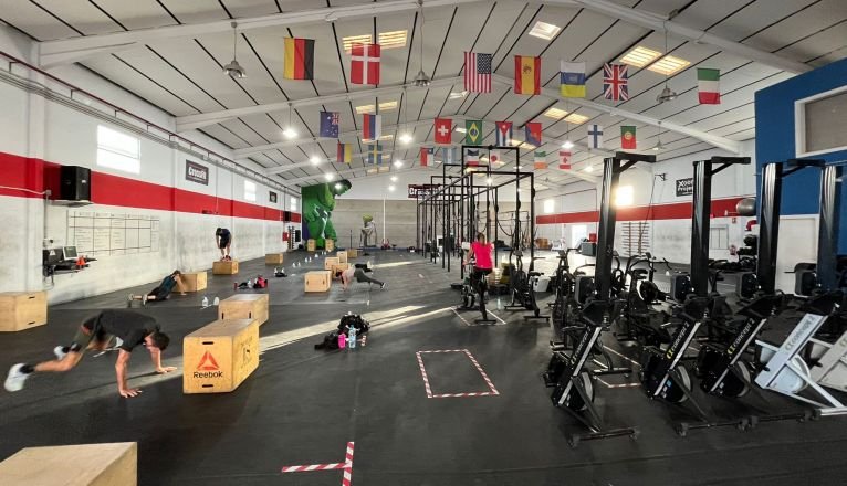 Ein Fitnessstudio mit verschiedenen Trainingsgeräten. Menschen trainieren mit Kisten und Geräten. Verschiedene Nationalflaggen hängen von der Decke. Im Vordergrund sind Rudergeräte zu sehen. Die Wände haben rote und weiße Streifen.