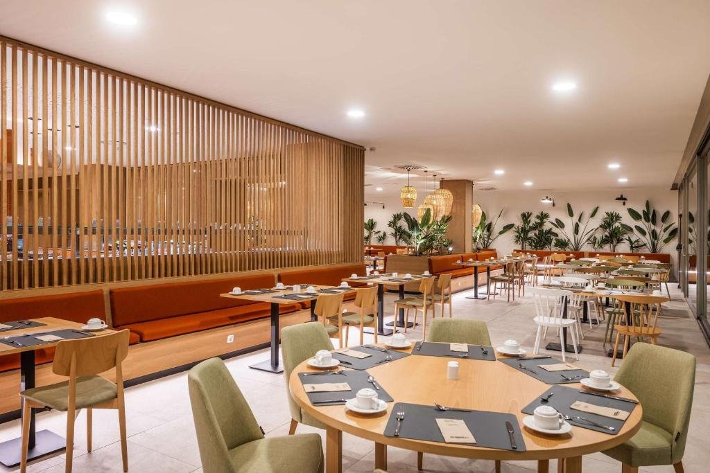 Ein modernes Restaurant mit Holztischen, grünen und holzfarbenen Stühlen und Pflanzen im Hintergrund. Die Tische sind mit Tellern, Tassen und Besteck gedeckt. Eine holzverkleidete Trennwand teilt den Raum.