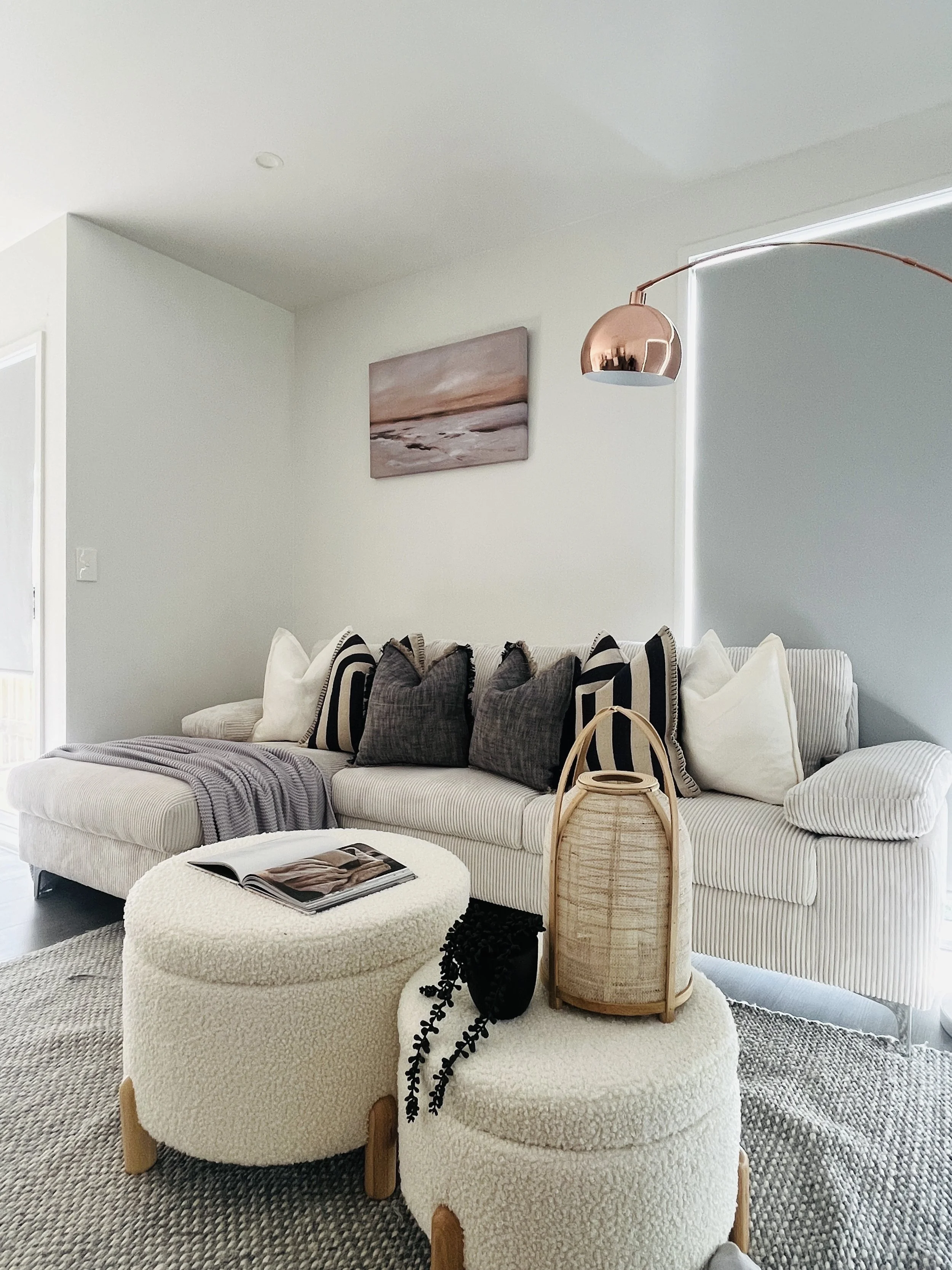 Modern living room with a light striped sofa, black and white pillows, a white textured ottoman with a magazine, a rattan lantern, black beaded decor, and a copper arc floor lamp. Artwork on the wall depicts a beach scene.
