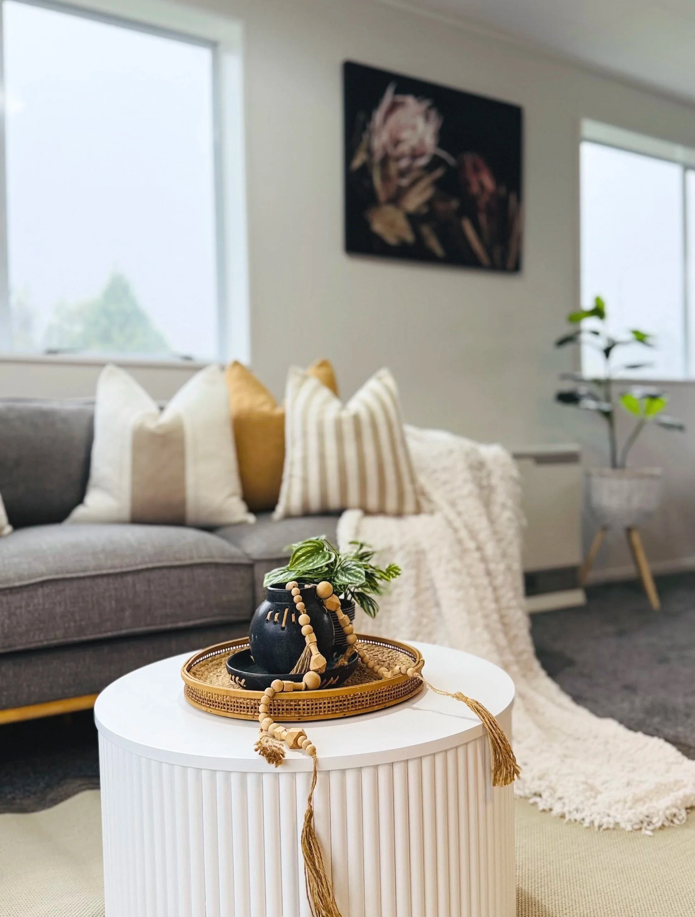 Living room with a gray sofa decorated with white and beige pillows, a white fluffy throw blanket, a black vase with green plants and wooden beads on a woven tray on a white round coffee table, a large window with natural light, a dark floral paintin