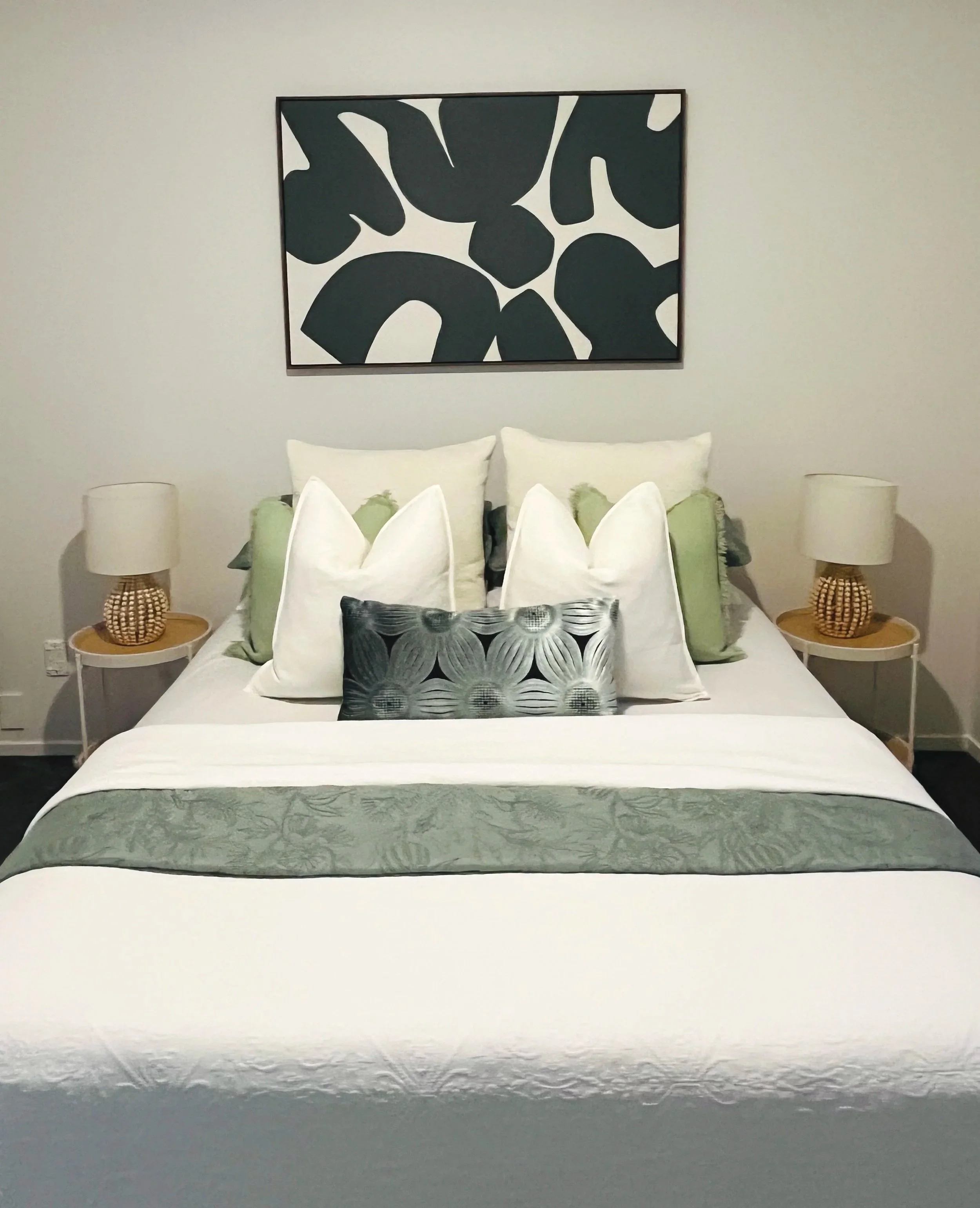 A neatly made bed with white bedding, multiple white and green pillows, and a gray floral runner. There are two matching white nightstands with wicker lamps on each side of the bed. An abstract painting with black shapes on a white background hangs a
