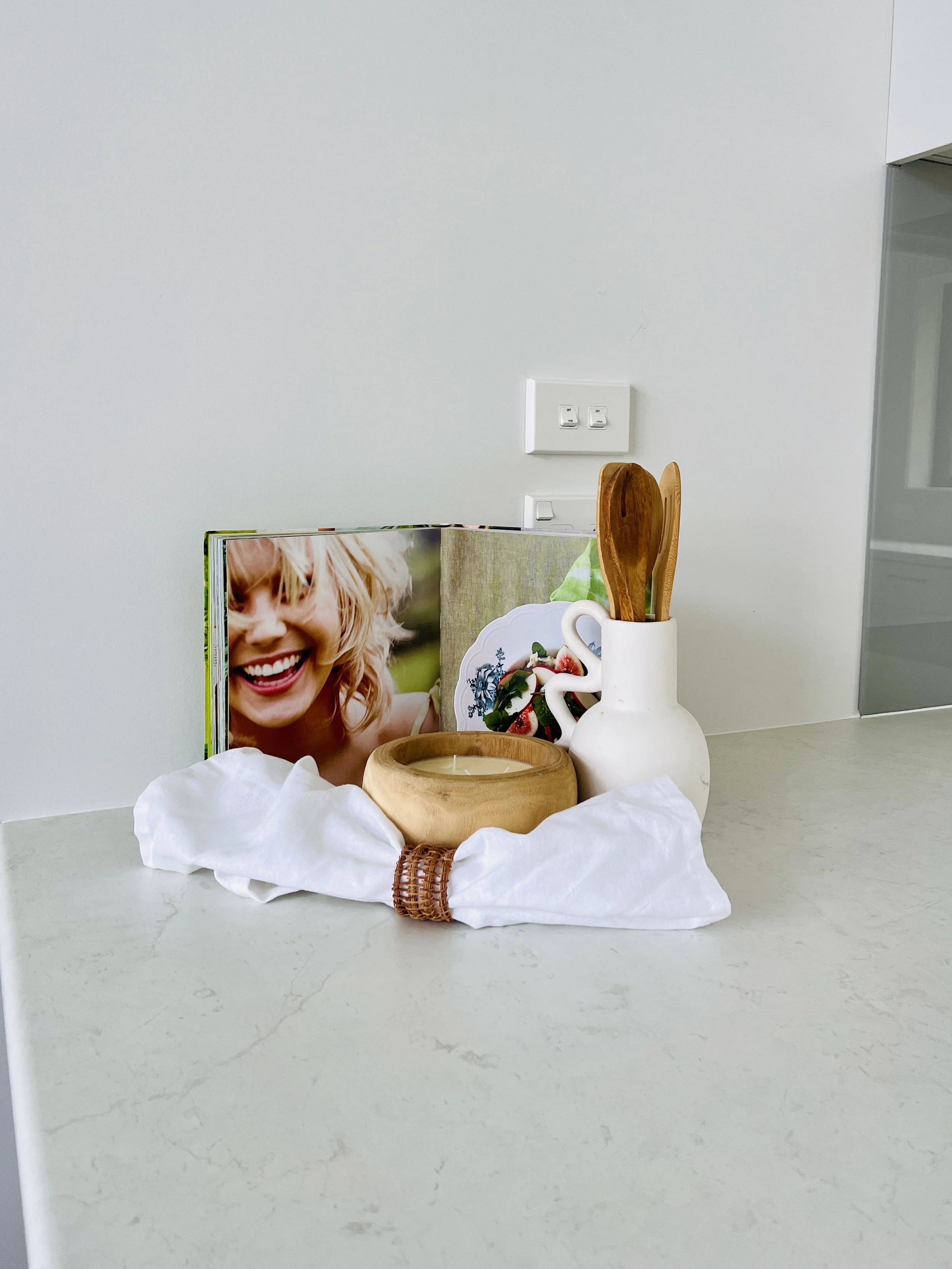 Decorative arrangement on a white surface, including a photo of a smiling blonde woman, a wooden bowl with a candle, a white ceramic pitcher holding wooden utensils, and a book or magazine with an image of strawberries and flowers.