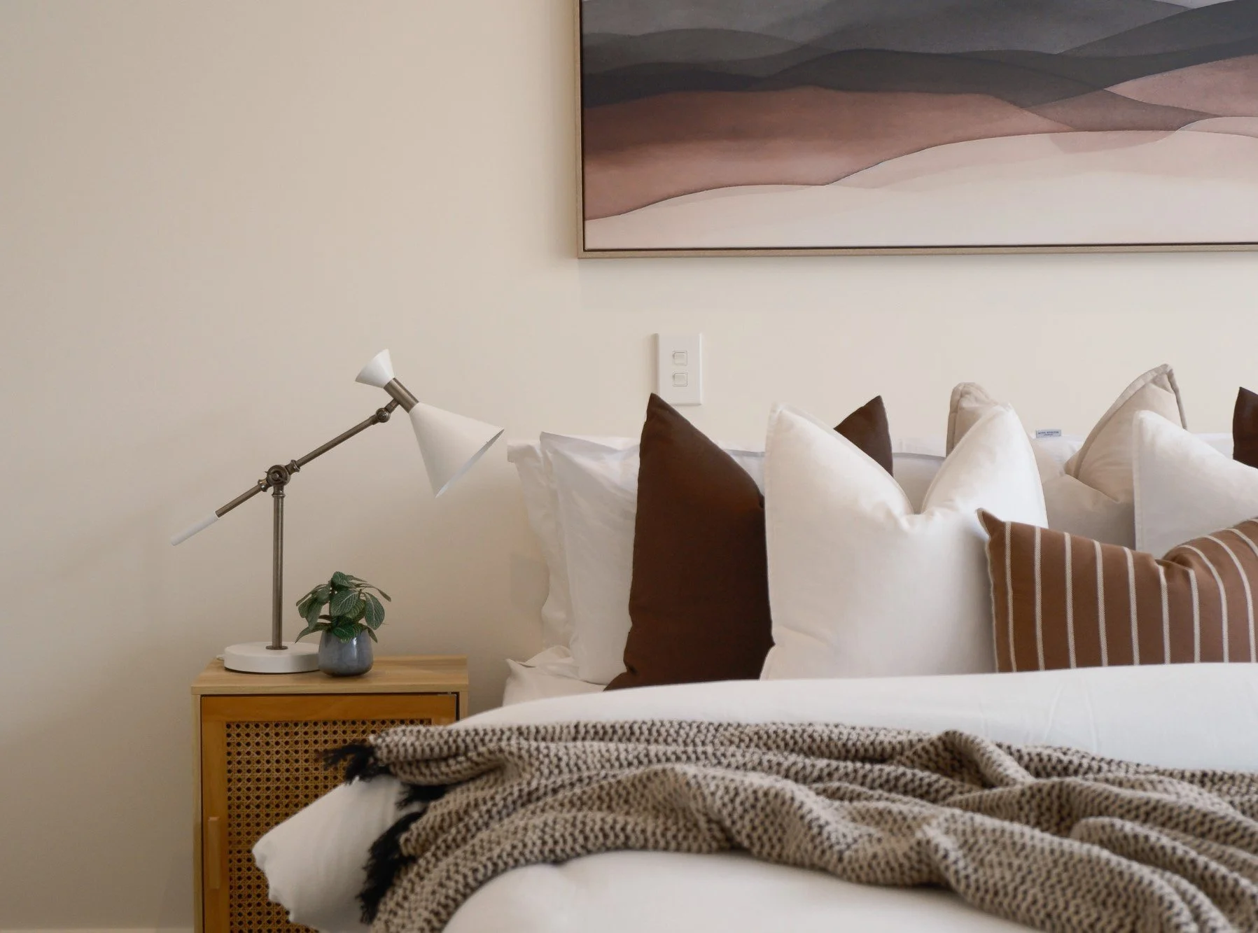 A cozy bedroom with a neatly made bed, multiple pillows in white, brown, and striped patterns, a beige blanket, and a wooden nightstand with a white adjustable desk lamp, a small potted plant, and a wall art piece with abstract landscape in soft eart