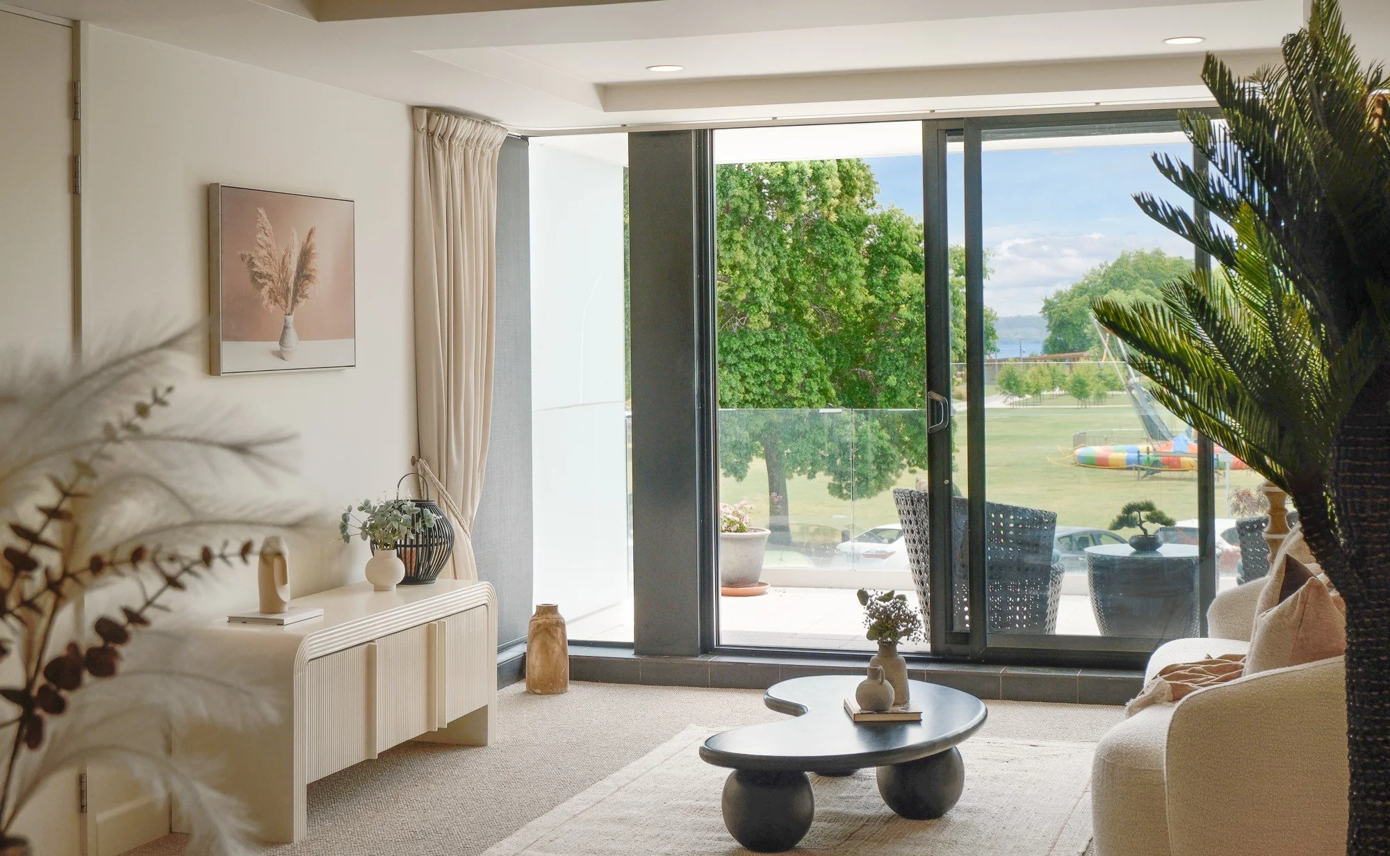 Living room with sliding glass door leading to balcony with outdoor seating and view of green park and water