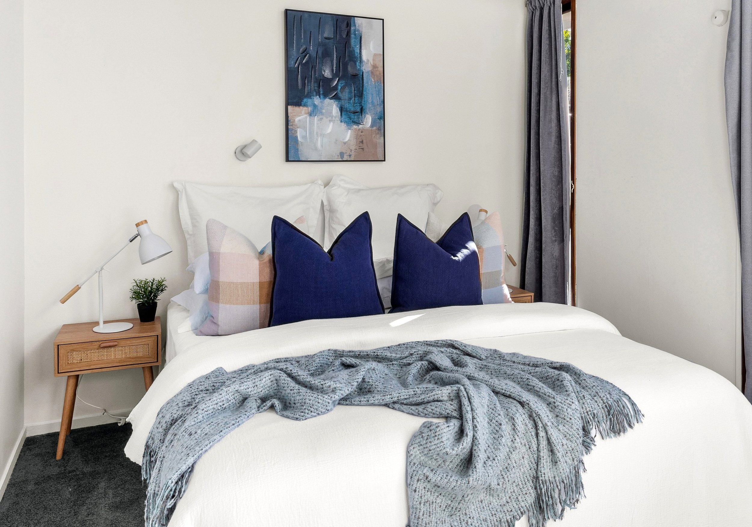 A neatly made bed with white bedding, two large white pillows, two decorative blue pillows, and two smaller plaid pillows. A gray knitted throw blanket is draped across the foot of the bed. On each side of the bed, there are wooden nightstands, with 